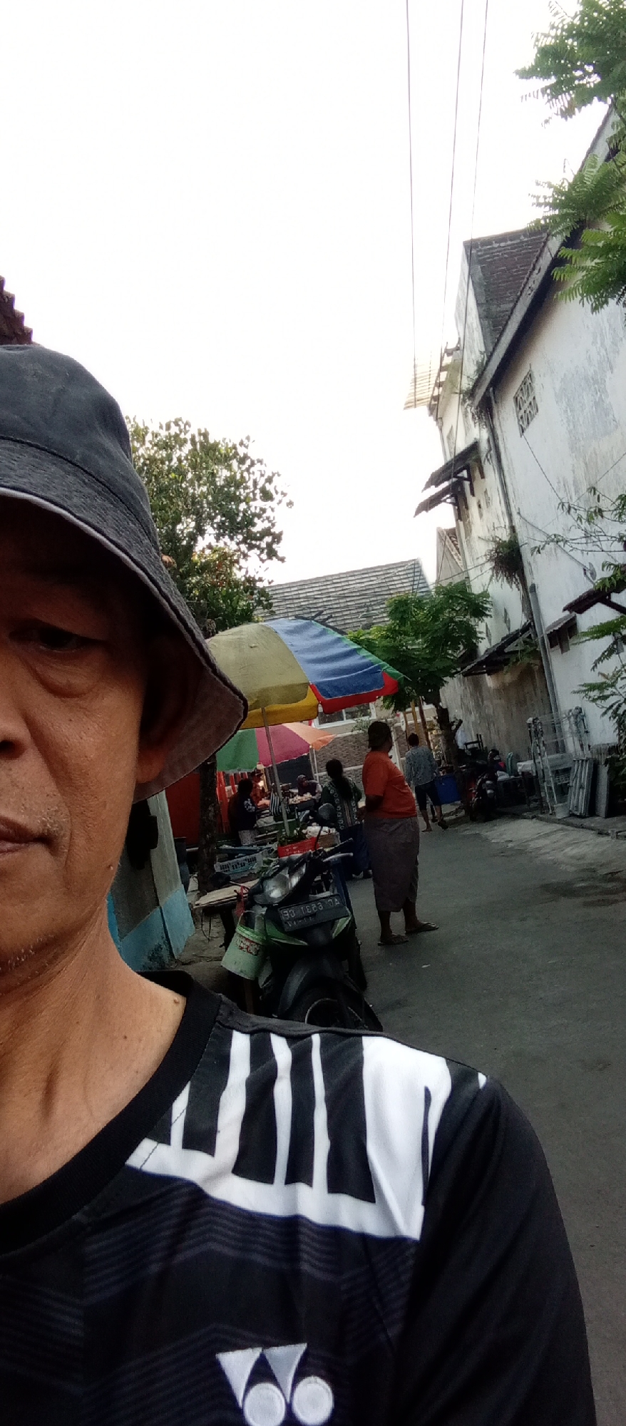 In the foreground, a person wearing a black bucket hat and a black and white striped t-shirt is visible. The t-shirt has a prominent logo at the bottom. In the background, a street scene unfolds with several elements. There are market stalls with colorful umbrellas, suggesting a market or food vendors. A green motorcycle is parked in the street. Several people are standing and walking around the market area. One person, wearing an orange shirt and a long skirt, is standing with their back to the camera. Another person in a checkered shirt is walking away from the camera. Buildings line one side of the street, with one building appearing to be multi-story with windows and balconies. Trees are visible in the background, and power lines cross the sky. The overall scene depicts a busy street in an urban environment, possibly in Kediri City, Indonesia, characterized by its market activity and residential buildings.