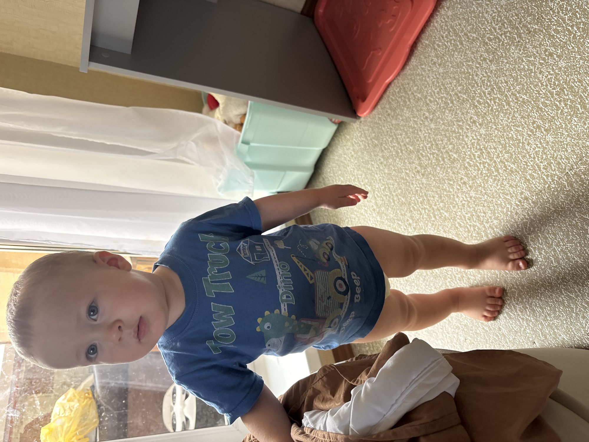 An indoor scene shows a toddler boy, approximately 12-18 months old, standing on a textured carpet. He has light brown hair and blue eyes, and is wearing a blue t-shirt with a "Tow Truck Dino" graphic and an orange diaper. The toddler is positioned towards the left side of the frame. To the left of the toddler is a brown blanket or cloth draped over furniture. In the background, there is a window with white curtains, a light blue plastic bin filled with stuffed animals, and a gray piece of furniture with a red tray on the floor next to it. The lighting appears to be natural, coming from the window. The scene does not provide any specific indicators to determine its location within the city of Naberezhnye Chelny, Russia.