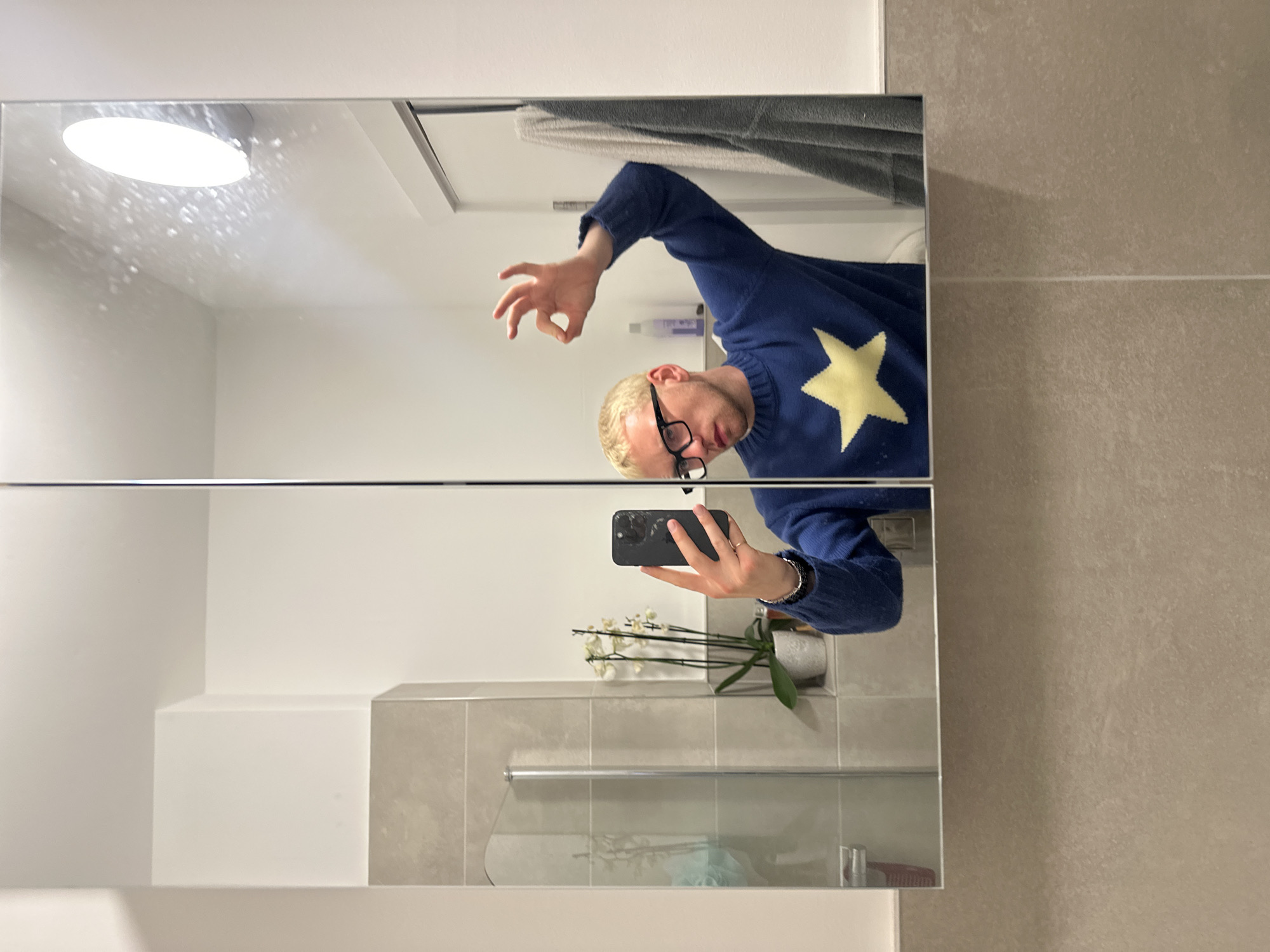 The image shows a mirror reflection of a person in a bathroom. The person has blonde hair, wears glasses, and is wearing a blue sweater with a large yellow star. They are holding a smartphone, presumably taking the photo. The bathroom has white and grey tiles, a light above the mirror, and a white orchid in a pot. The image provides no information about the city of London.