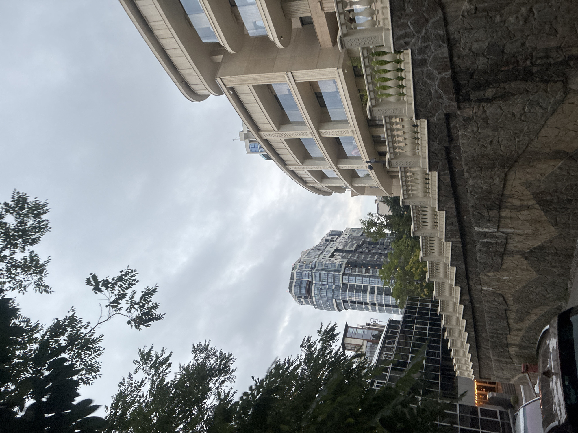 The image depicts a modern urban landscape with a cloudy sky overhead. In the foreground, a tiered stone wall is visible, topped with a white ornate balustrade that follows a curved path. To the right of the stone wall, a multi-story curved building with balconies and large windows is prominent. Behind this curved building and to the left, a tall, modern skyscraper with a distinctive blue-tinted glass facade stands out. In front of the skyscraper, a lower building with a dark glass facade and some illuminated windows is partially visible. Trees with dark green foliage frame the scene from the left and partially obscure the sky. The overall impression is one of contemporary architecture against a muted, overcast sky, suggesting a city setting in Sochi, Russia. There are no people visible in the image.