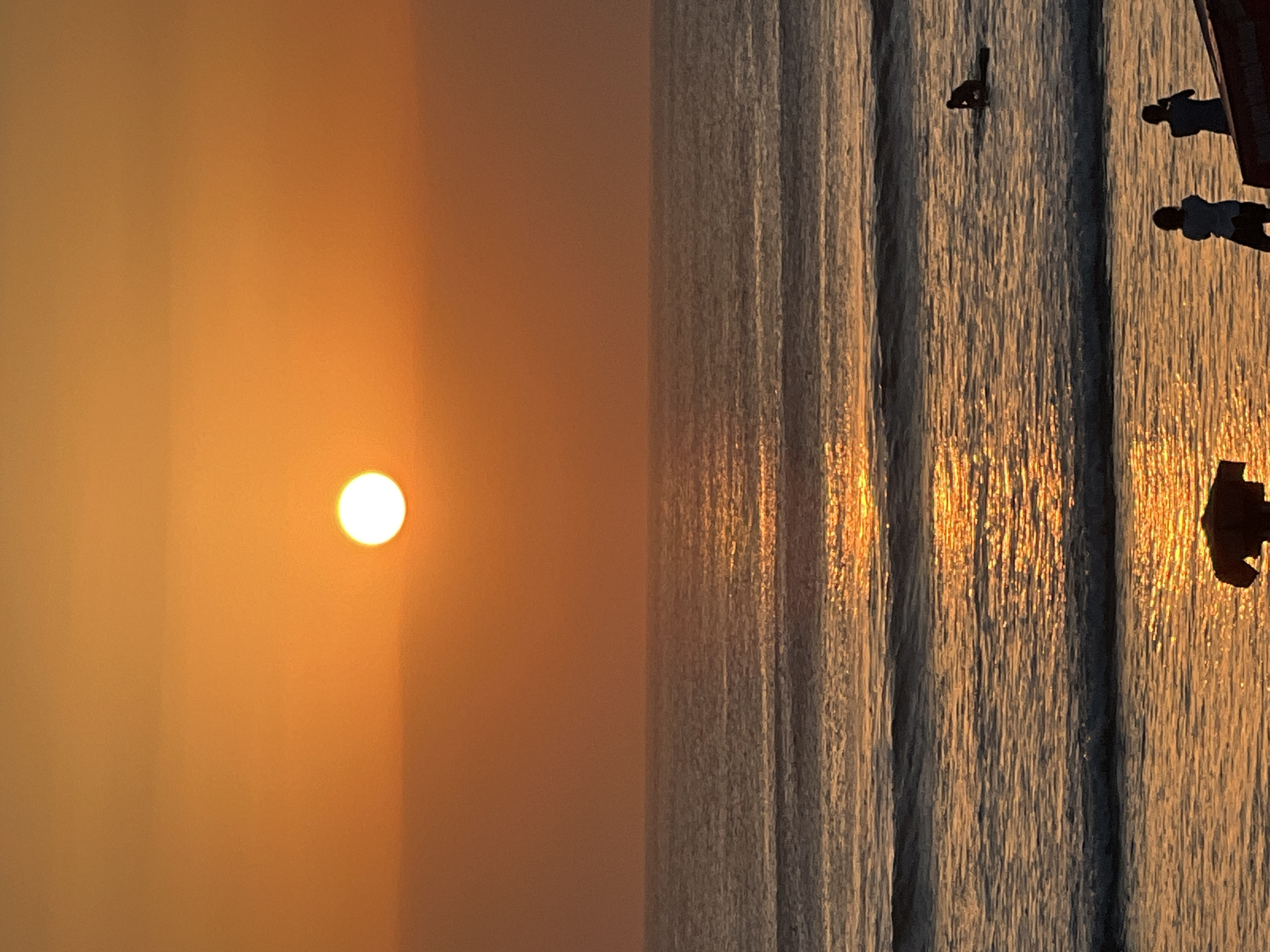 This image depicts a serene sunset scene at Kuta Beach, Indonesia. The sun, a bright orange orb, hangs low in the sky, casting a warm, golden glow across the water and the horizon. Gentle waves roll towards the shore, their surfaces shimmering with the reflected light of the setting sun.
In the foreground, two figures are silhouetted against the vibrant sunset, standing near the water's edge. Their postures suggest they are observing the sunset or perhaps engaged in a conversation. Further out in the ocean, a lone surfer is visible on a surfboard, seemingly taking a break or waiting for a wave. The ocean stretches to the horizon, where it meets the soft, gradient hues of the sky, ranging from a deep orange near the sun to lighter tones as it ascends. The overall atmosphere is one of tranquility and natural beauty.