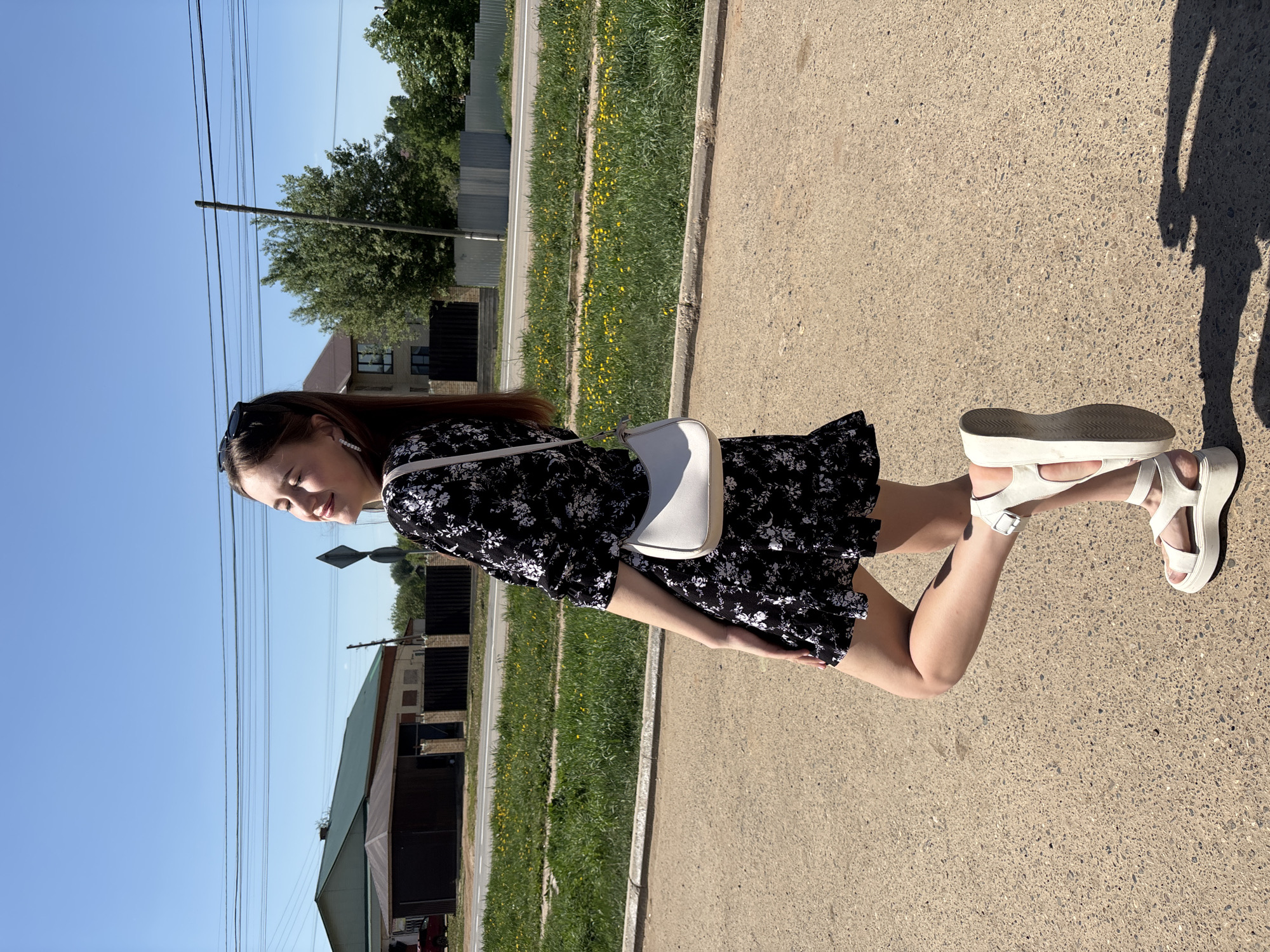 A young woman stands on a paved area, possibly a sidewalk or road, in a location identified as Фоки, Russia. She wears a short black floral dress, white platform sandals, and a small white shoulder bag. She is looking to her left with a slight smile. The background includes a mix of residential and possibly industrial buildings, power lines against a clear blue sky, and patches of green grass and yellow flowers along the roadside. The image is taken during the daytime, and a shadow is visible on the paved surface.