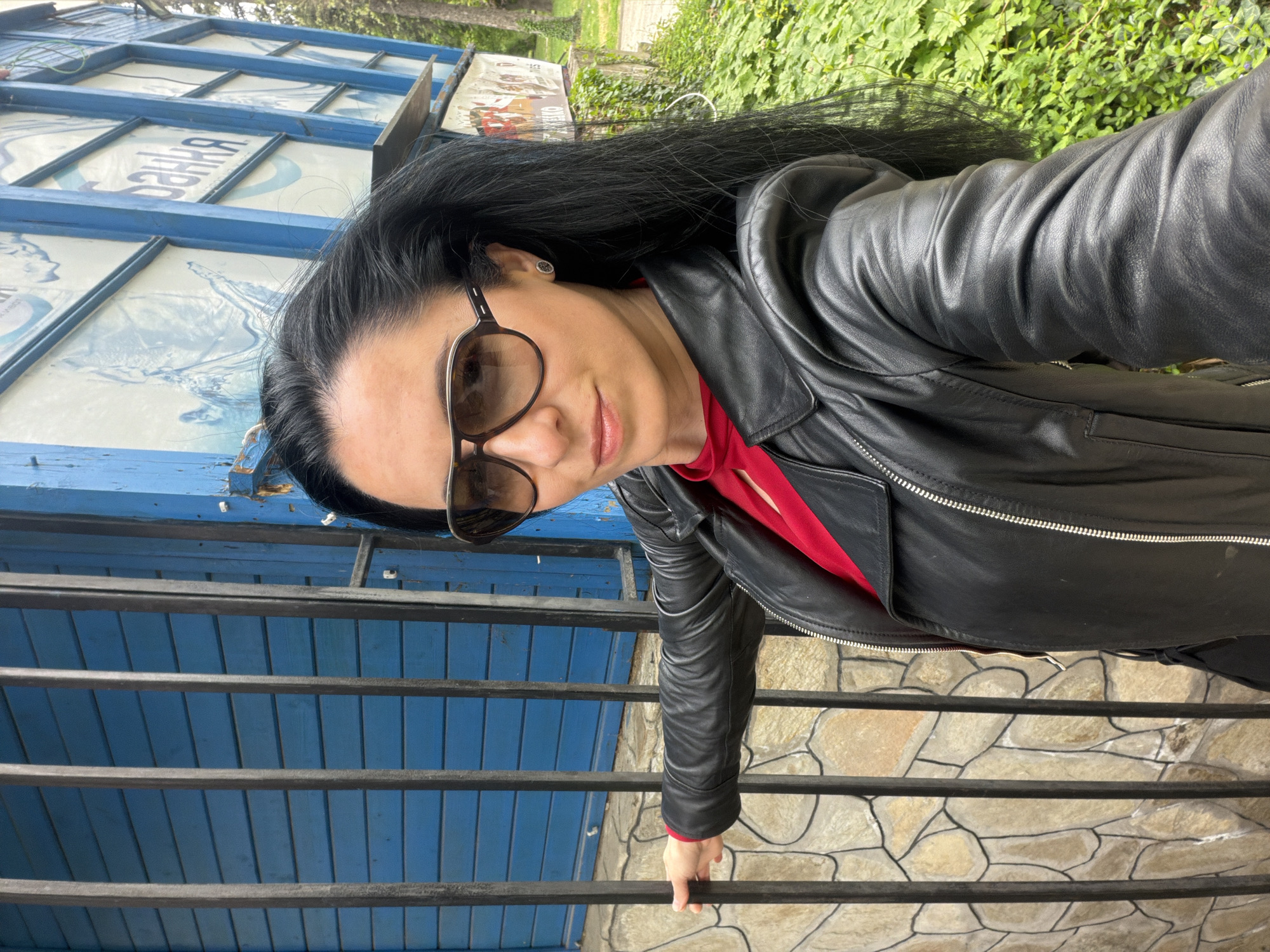 The image depicts a woman with long black hair, wearing sunglasses and a black leather jacket over a red top. She is standing in front of a structure with blue and white signage and a stone wall. The structure has metal bars. The location is likely in the city of Pleven, Bulgaria.
