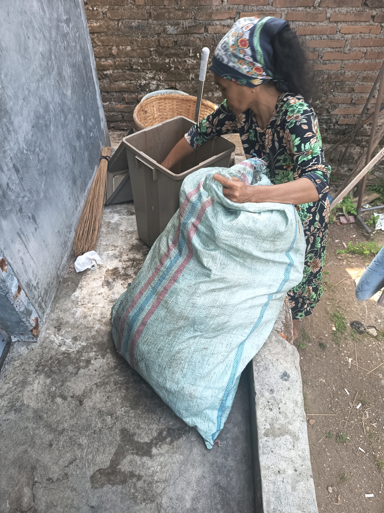 In a narrow alleyway between two brick walls, a woman wearing a floral-patterned dress and a headscarf is actively engaged in filling a large, light blue woven sack with material from a dark gray plastic bin. Beside the bin, a broom made of natural fibers leans against a light gray wall. The ground is a mix of concrete and dirt. In the background, a wicker basket is visible, and to the right, there are some wooden planks and scattered debris. The setting appears to be an outdoor area in the city of Banjar, Indonesia.