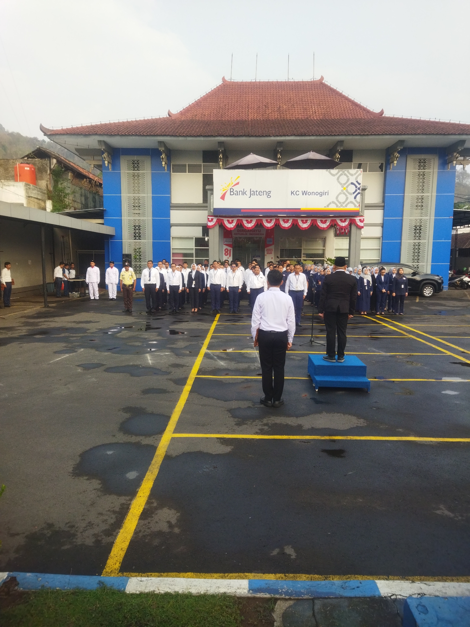The image depicts an outdoor gathering in front of a building identified as "Bank Jateng KC Wonogiri" in Giripurwo, Indonesia. A group of people, predominantly wearing white shirts and dark pants or skirts, are assembled in a parking lot, suggesting a formal occasion like a flag-raising ceremony or assembly. Some individuals are standing in formation, while others appear to be observing. In the foreground, a person in a white shirt and black pants stands facing a group of people, positioned on a small blue platform. Another individual in a black suit stands on the platform, facing away from the camera and towards the assembled group. The building's architecture features a traditional Indonesian-style roof with red tiles and a blue facade. The ground is asphalt, marked with yellow lines, and shows signs of recent rain with puddles visible. In the background, a dark-colored car is parked. The overall atmosphere suggests an organized event taking place in a semi-urban or institutional setting.