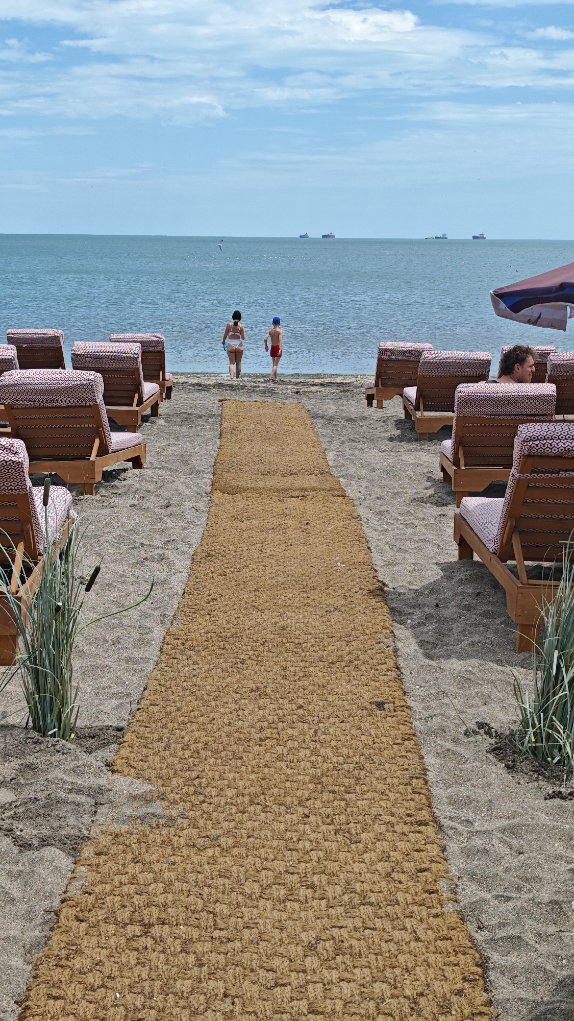 The image shows a beach scene, likely in Burgas, Bulgaria, given the location's association with seaside resorts. In the foreground, a woven mat path stretches towards the sea, flanked by beach chairs arranged in rows. The chairs feature a patterned fabric upholstery and are constructed with wooden frames. Vegetation grows sparsely on either side of the path, including tall grasses and cattails. Two figures, an adult woman in a white bathing suit and a young boy in red swimwear and a blue cap, are walking towards the water at the end of the path. In the background, the sea horizon is visible, with several ships silhouetted against the light-blue sky. The sky is mostly clear with a few scattered clouds. A partially visible beach umbrella and a reclining man are seen amongst the beach chairs to the right of the path. The sand appears dark and coarse.