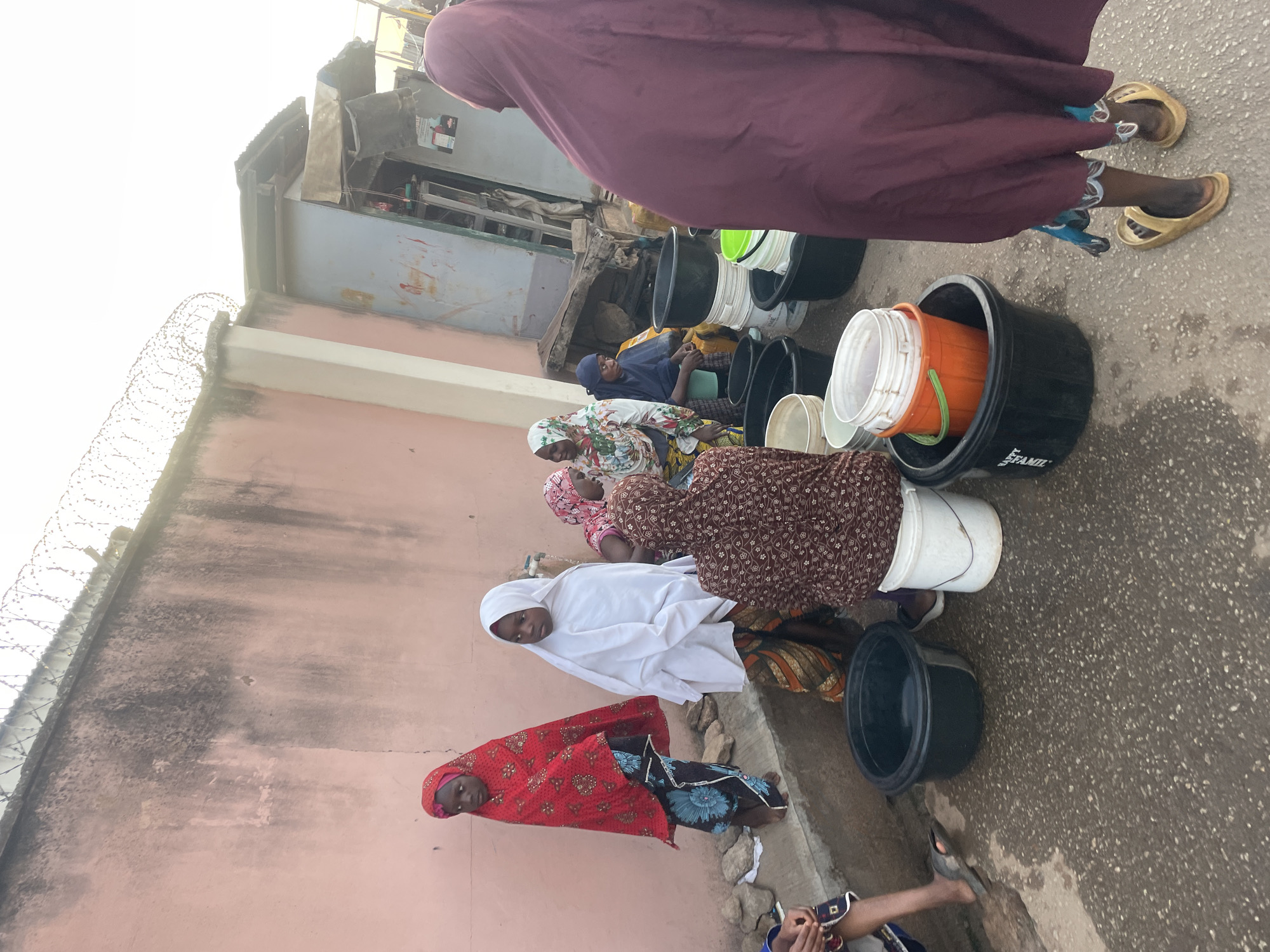 In Jos, Nigeria, a line of women sit on the ground next to large black buckets and smaller white or orange buckets. They are outside a building with a pink wall topped with barbed wire. The women are wearing long, colorful dresses and headscarves. One woman is wearing a red headscarf and a blue and black dress, another is in a white headscarf. They seem to be waiting in line, possibly to collect water. The ground is paved and there is another building in the background.