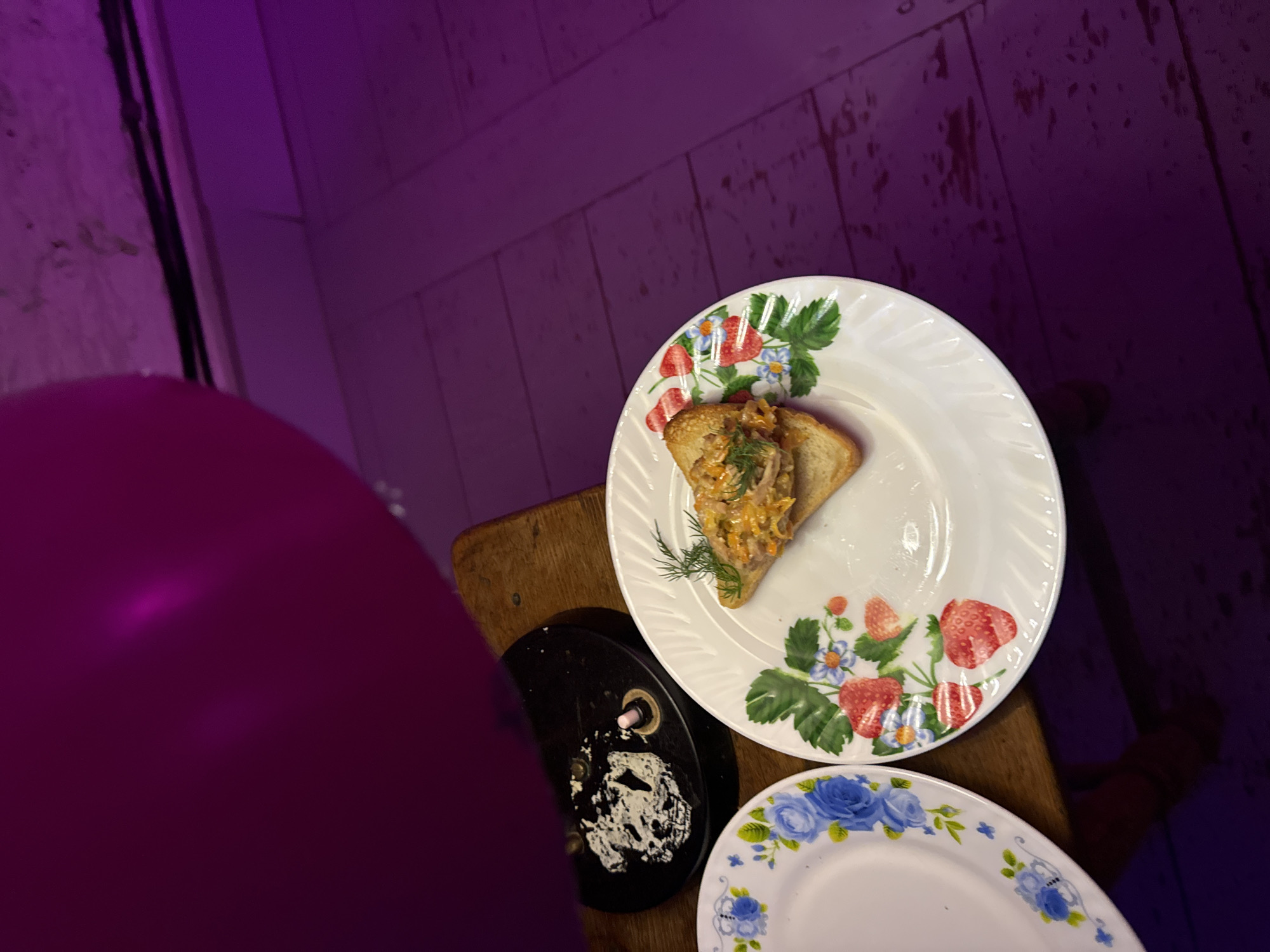 The scene presents a top-down view of a dining setting, likely indoors, characterized by a mix of domestic and culinary elements. A plate with a white base, adorned with a strawberry and floral pattern, is prominently featured. On it rests a piece of toast topped with what appears to be some kind of spread. Beside this plate is another plate. The setting includes a dark, possibly soiled, surface adjacent to the plate, along with a portion of a wooden surface. A large pink or magenta-colored balloon occupies a significant portion of the foreground. The backdrop consists of a planked surface, possibly a wall. The scene is lit with a purple hue. The location is not discernable from the image.