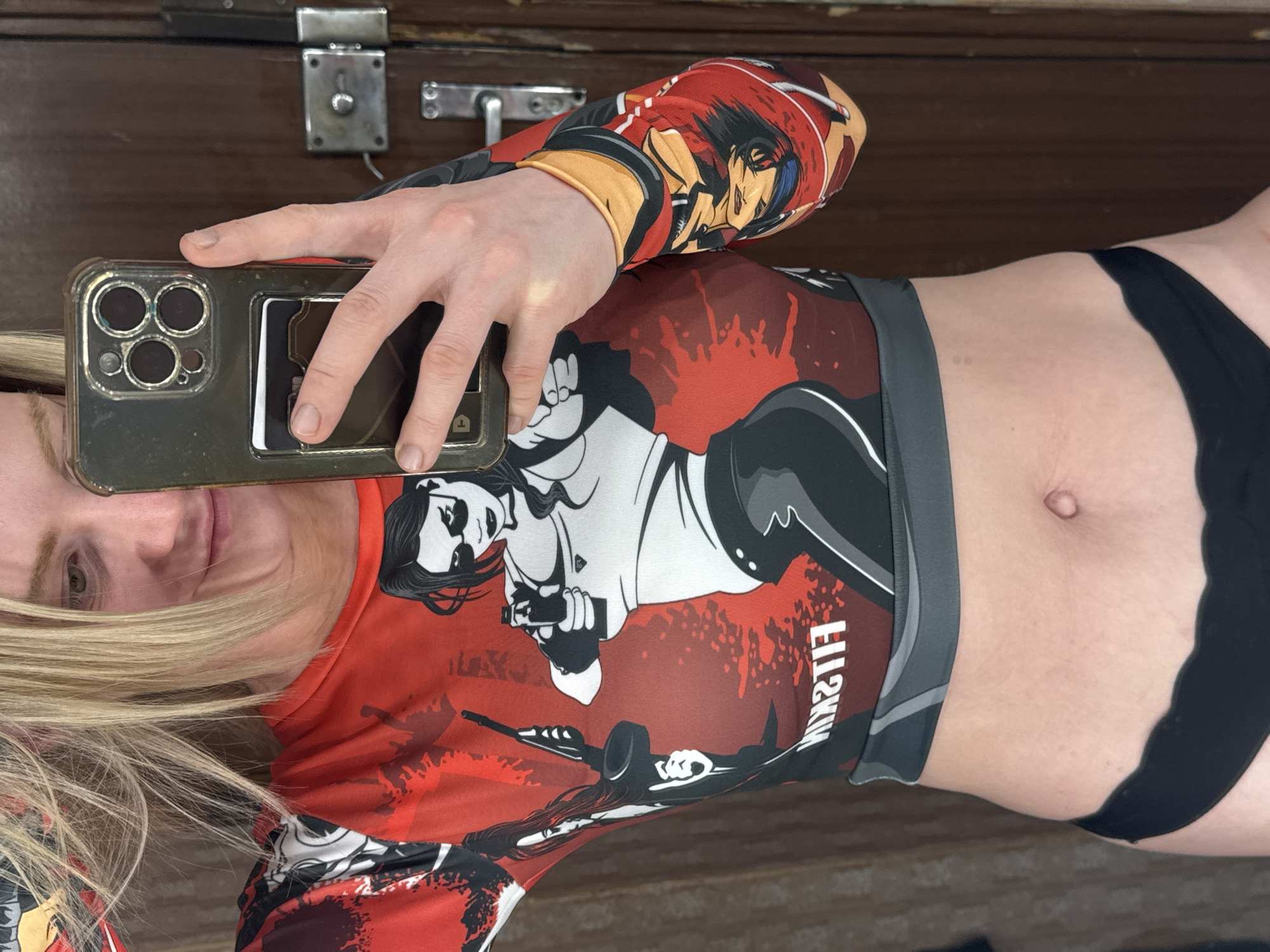 A female is taking a selfie in front of a wooden door with silver hardware. She is wearing a red, grey, black, and white rash guard with images of women holding weapons and characters from an anime. She is also wearing black underwear. Her hair is blonde. She is holding a cell phone in her left hand, which is covering part of her face.
I cannot determine the location based on the image.