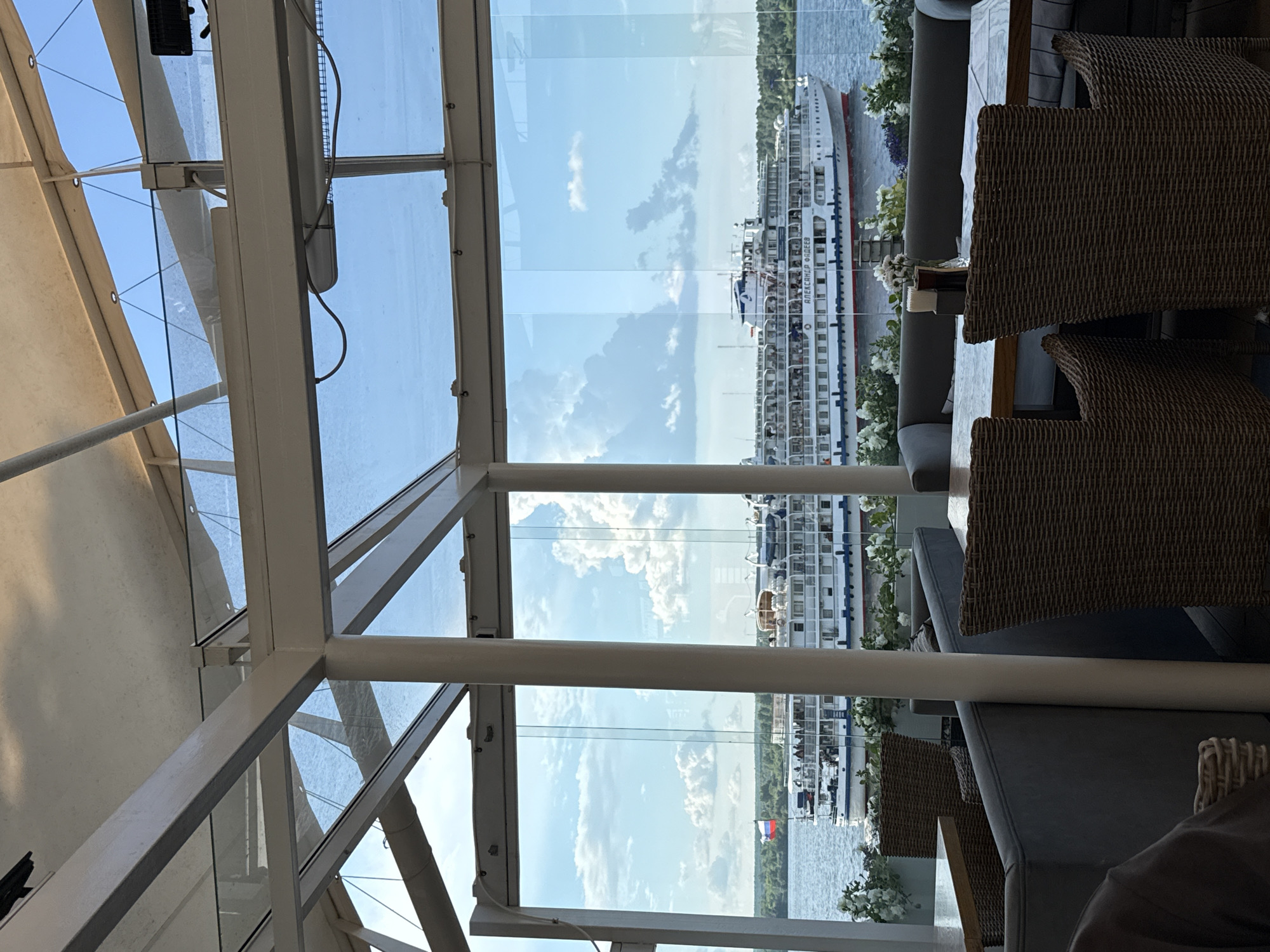 The image captures a view from an indoor seating area, likely a restaurant or cafe, with large glass windows offering a scenic outlook. Through the windows, a large white cruise ship with multiple decks is visible on a body of water. The ship has the name "АЛЕКСАНДР ФАДЕЕВ" written on its side. A Russian flag flies on the left side of the ship. The background consists of a green treeline and a sky filled with white clouds. In the foreground, inside the seating area, there are tables with wicker chairs and upholstered benches, suggesting a dining or lounge setting. The architectural elements of the indoor space, including beams and a glass ceiling, are also visible. This scene is likely the restaurant "Severnye Tipy" (North Types) in Perm, Russia, which is known for its views of the Kama River and riverboats.