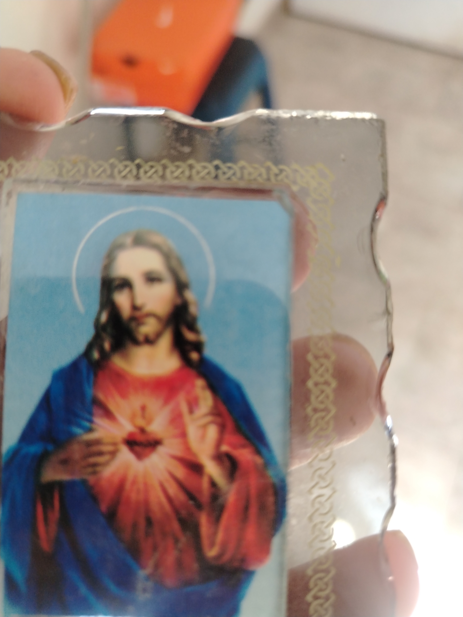 The image shows a close-up of a religious icon, likely a small framed image of Jesus Christ. The icon depicts Jesus with a halo, long hair, and a beard. He is wearing a blue robe over a red garment, with his right hand placed over his chest where a radiant heart is visible. The icon is framed by a clear, slightly beveled material with a decorative yellow border. A finger and thumb, holding the icon, are partially visible on the left side of the frame. The background is out of focus, showing what appears to be an indoor setting with an orange object and a dark blue object in the background. The lighting suggests a bright environment, possibly with natural light from the right. There is no specific indication of the location in Juan Griego, Venezuela, within the image itself.