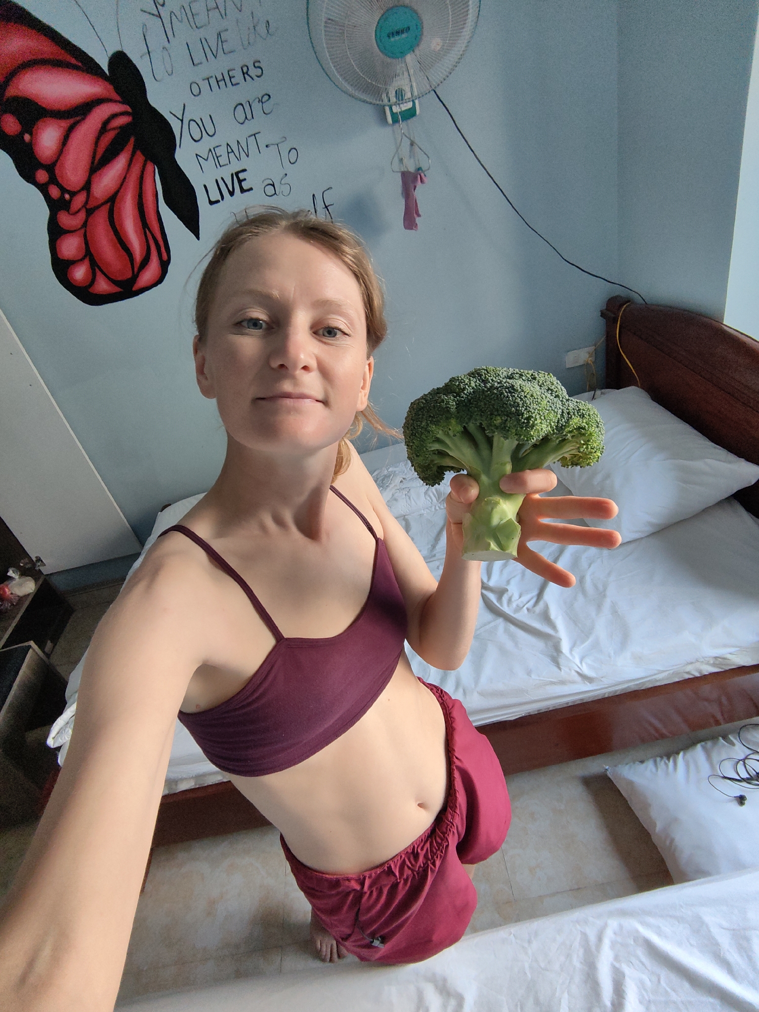 A woman stands in a bedroom, holding a head of broccoli in her right hand. She is wearing a maroon sports bra and maroon shorts. A wooden bed with white sheets is behind her, and a black and red butterfly mural is on the light blue wall to her left. A white fan is mounted on the wall above the head of the bed. The floor is tiled. The location is Bai Chay Ward, Vietnam.
