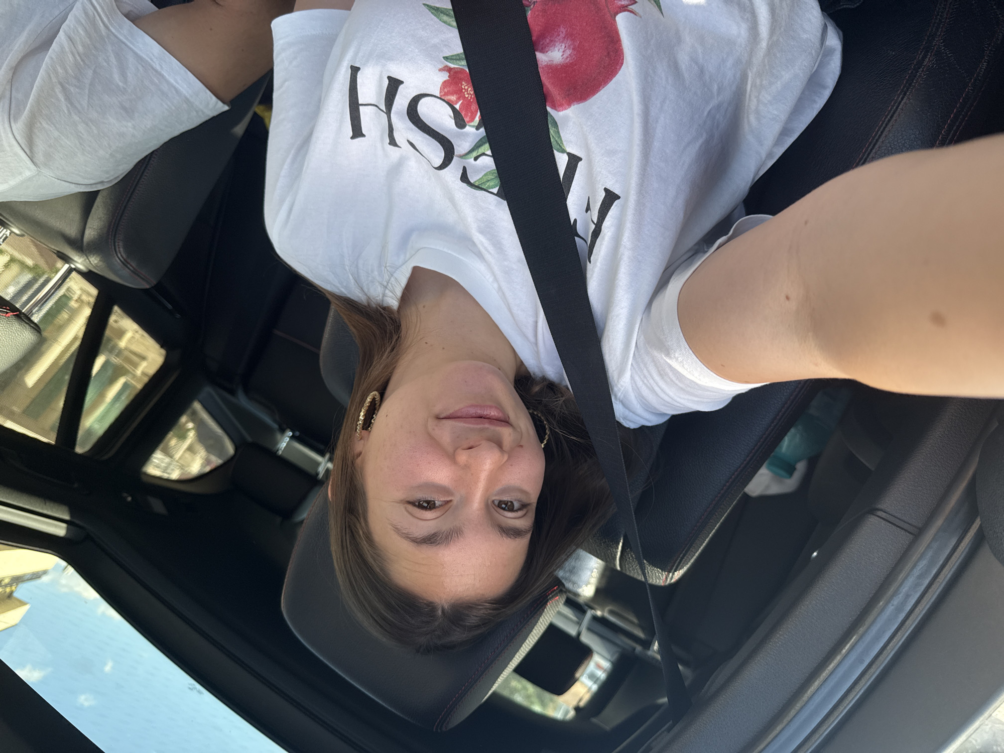 A young woman with brown hair and large hoop earrings is taking a selfie in the passenger seat of a car. She is wearing a white t-shirt with a graphic of pomegranates and the word "FRESH" printed on it. A black seatbelt is diagonally across her chest. The interior of the car is visible, including the black leather seats and a sunroof. The image appears to have been taken in Sofia, Bulgaria, due to the presence of a water bottle with a blue cap, commonly found in Bulgarian stores, and the bright sunlight suggesting an outdoor environment.