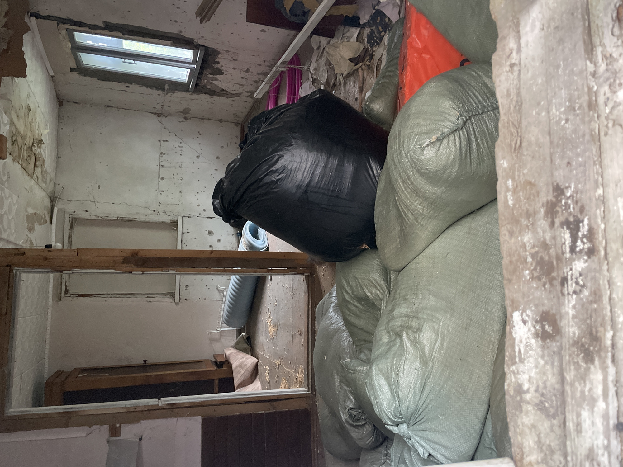 An interior view of a dilapidated structure, possibly a room or storage area, filled with large, tied-off sacks of indeterminate contents, mostly olive green. One black bag is visible amidst the green ones. An orange bag is also partially visible. The bags are blocking part of the entrance and take up most of the space. Part of another room or area is visible through a rectangular opening or window with a wood frame, showing more of the interior and a wooden cabinet. The walls are peeling and damaged, the ceiling shows water stains and damage. There's a window near the ceiling. The location is Olginka, Russia. No people are visible in the image.