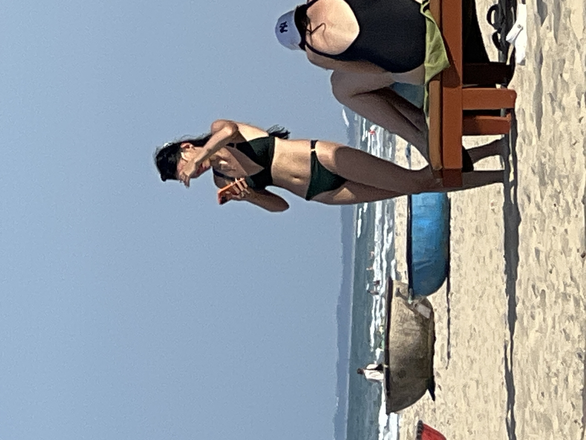 The image shows a beach scene in Da Nang, Vietnam. In the foreground, two women are visible. One woman, wearing a dark green bikini, is standing and holding a smartphone, shielding her eyes from the sun with her hand. The other woman is seated on a wooden lounge chair, facing away from the camera, wearing a dark swimsuit and a light-colored baseball cap. Various objects are scattered on the sand, including a blue bucket and what appear to be fishing boats. In the background, the ocean is visible with waves and several people swimming or wading in the water. The sky is clear and blue.