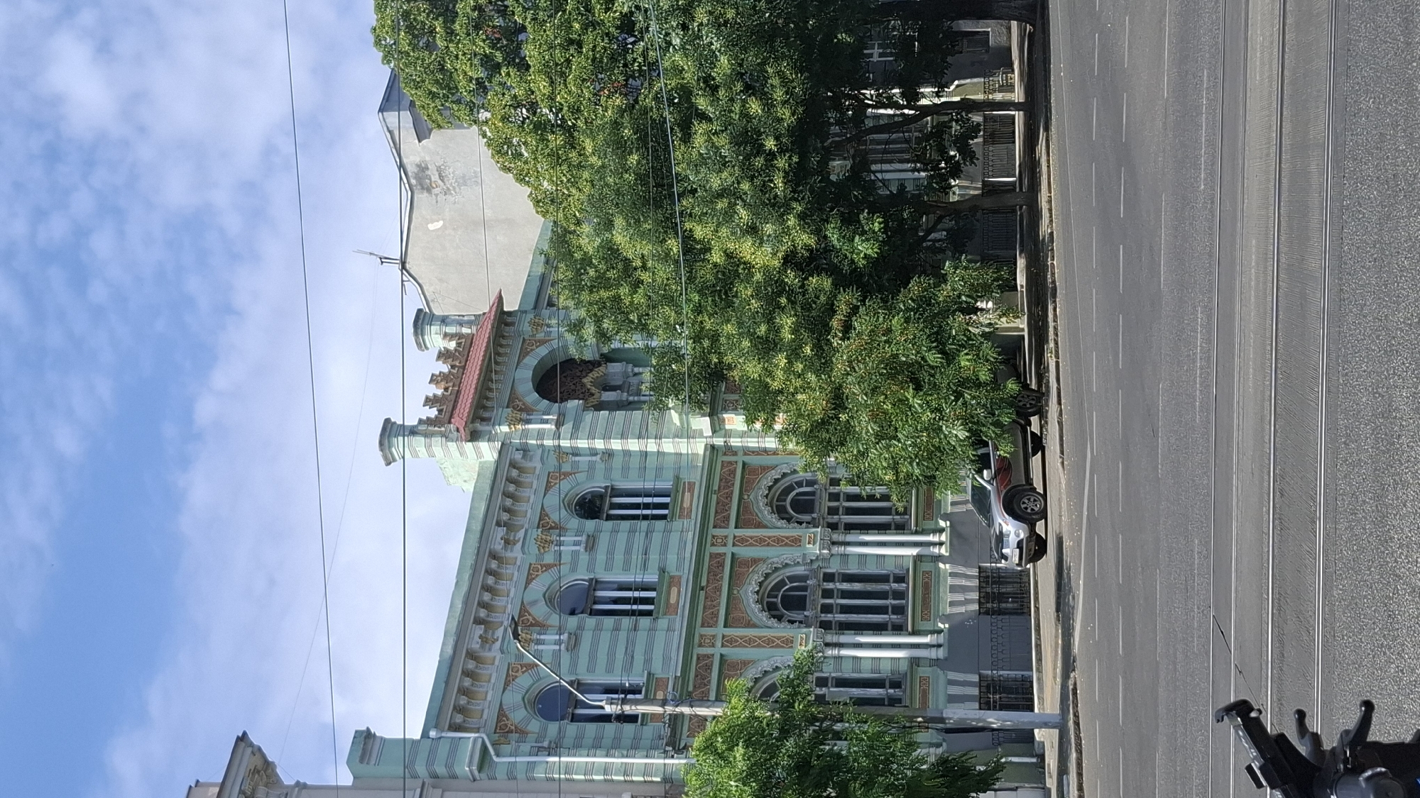 An exterior, eye-level, medium shot captures a light-green historic building with elaborate ornamentation, set against a clear blue sky with scattered white clouds. The building features arched windows, decorative friezes, and a red-tiled roof section. In front of the building, a silver SUV with red decals is parked on a street. Lush green trees partially obscure the lower sections of the building and line the sidewalk. The foreground shows a paved road with white lane markings and tram tracks. The overall scene depicts a street view in an urban environment.