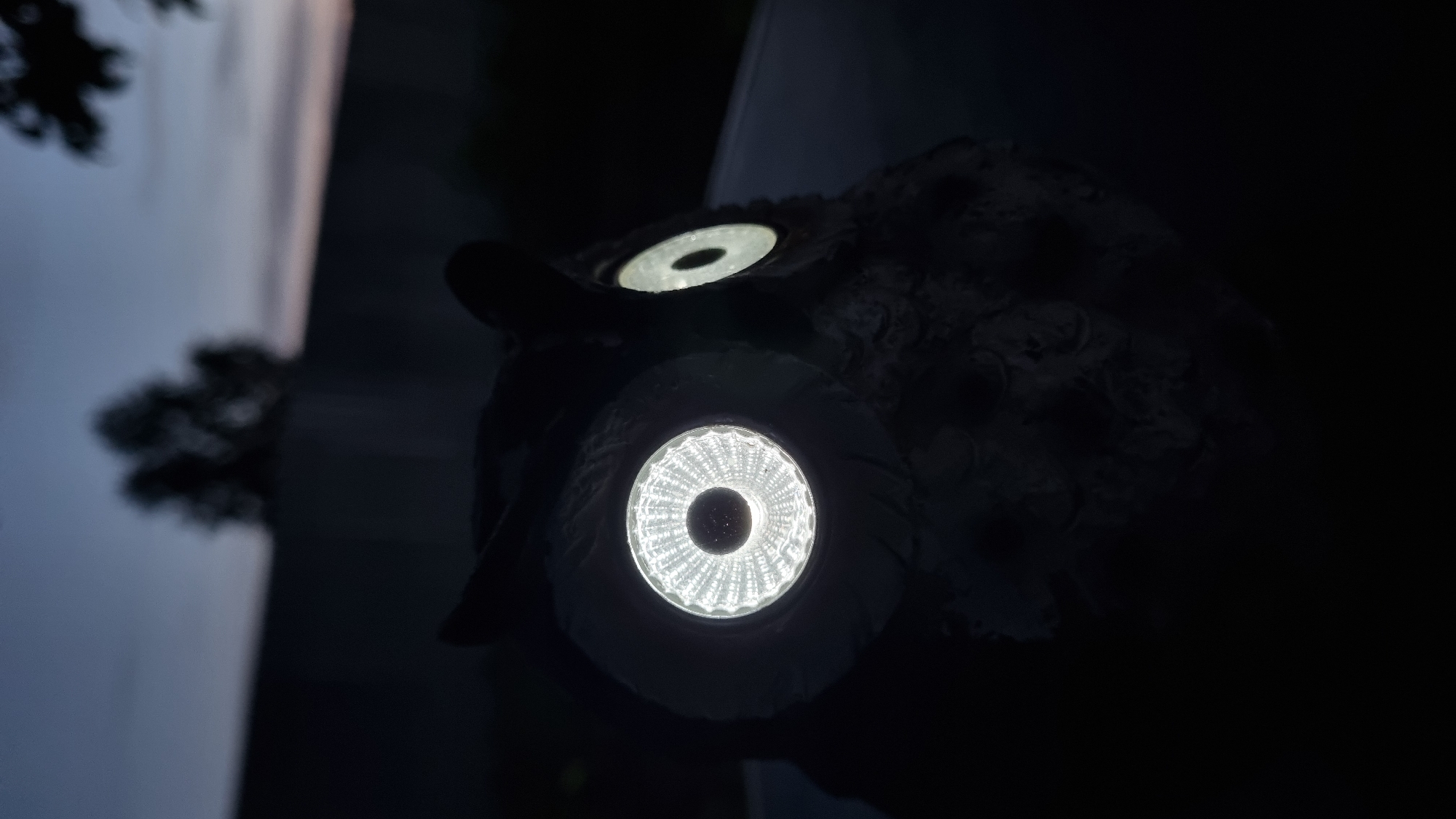 An owl-shaped garden ornament with glowing eyes is positioned outdoors at dusk. The owl is dark gray and appears to be made of stone or resin. Its large, round eyes are illuminated with bright white light, with the left eye showing a more textured pattern. The ornament is placed on a dark surface, possibly a table or railing, with a blurred background of a fence and trees against a twilight sky. The image captures a serene and slightly mysterious atmosphere. The location is the city of Vysokoe, Russia.