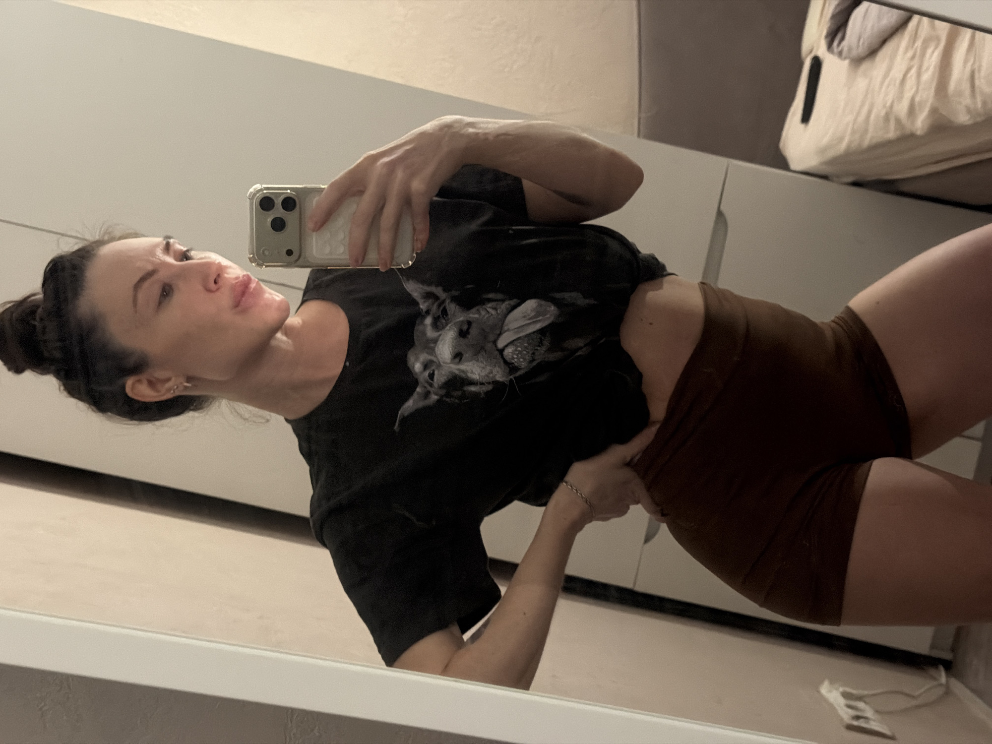 A woman is taking a selfie in front of a mirror. She has dark hair pulled back in a bun. She is wearing a black t-shirt with a dog graphic and brown shorts. The mirror reflects a room with a white cabinet or wardrobe, and a bed with a blanket in the background. The electrical outlet is visible on the wall. The location is likely an apartment or residential space. There is no evidence of it being in Perm, Russia.