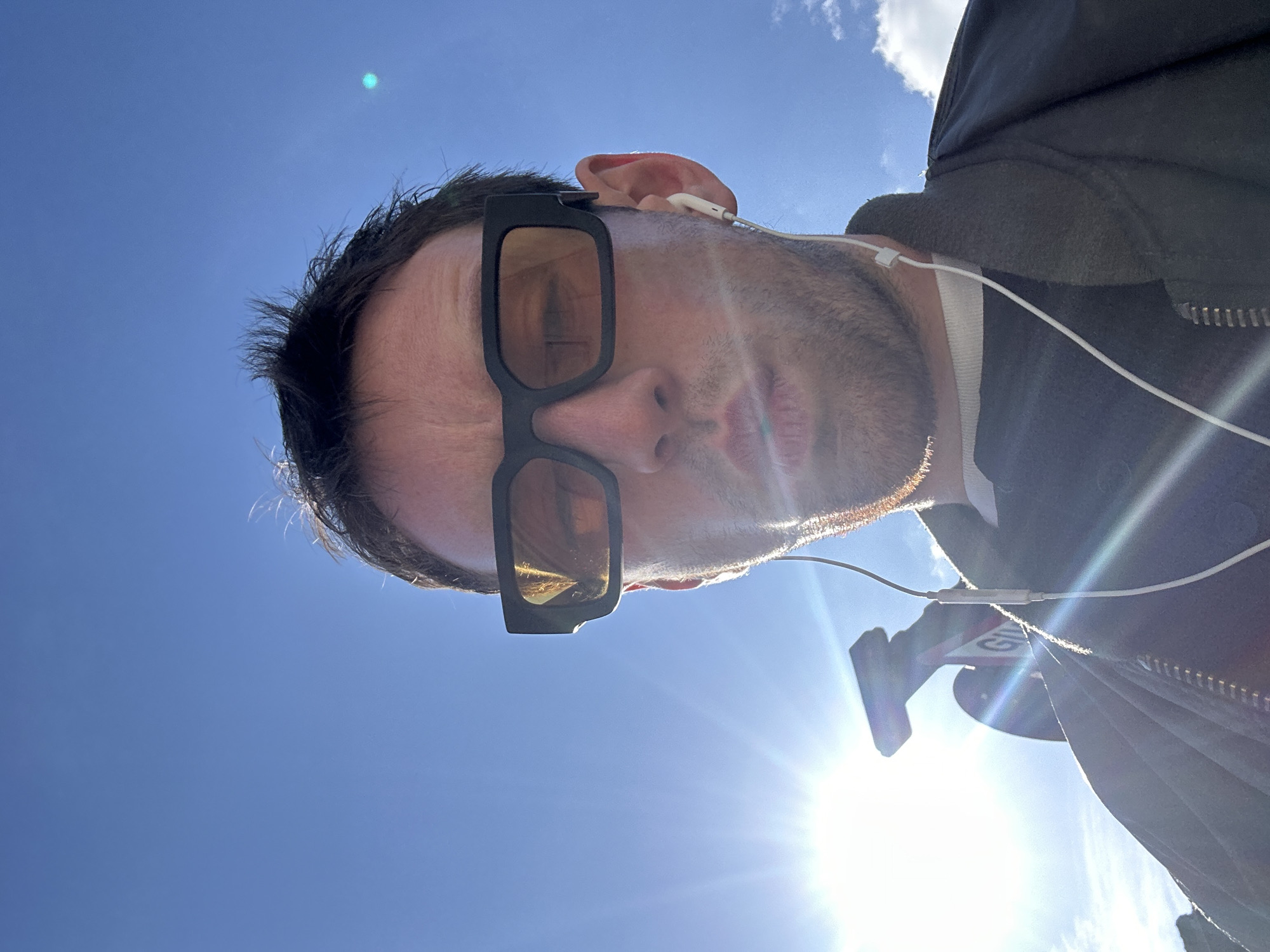 A selfie shot from a low angle looking up at a Caucasian male wearing sunglasses and earphones, taken on a sunny day. The background features a clear blue sky. The man is wearing a dark-colored jacket. Part of a sign is visible near his shoulder. The location is the city of London, United Kingdom.