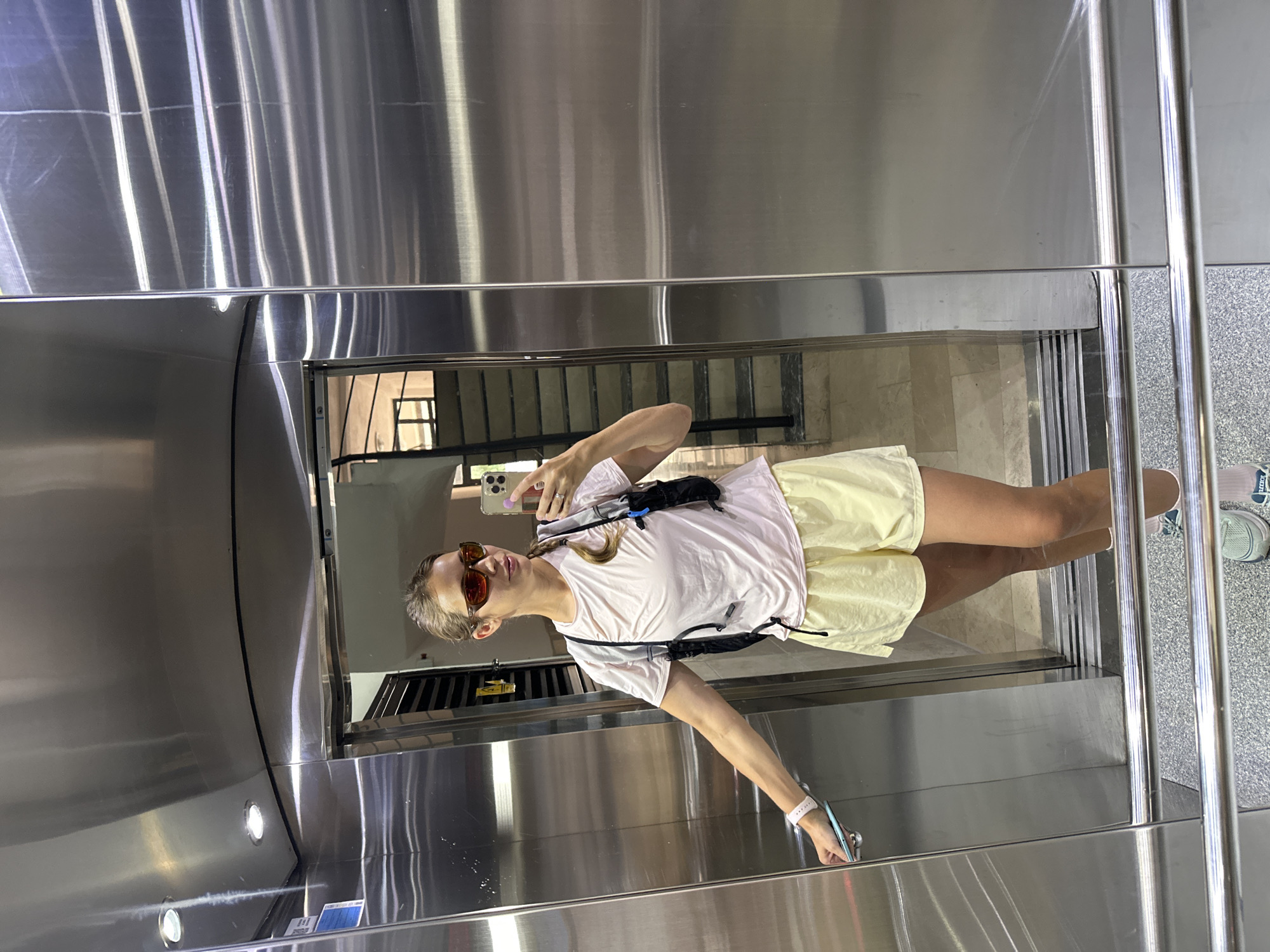 A woman takes a selfie within a modern, highly reflective metallic elevator, likely situated in Fethiye, Turkey. The image, oriented horizontally, shows the woman's reflection as she holds a smartphone (resembling an iPhone) in her right hand, pointed towards the mirrored surface. She wears dark sunglasses, a light pink short-sleeved t-shirt, and light yellow shorts. A black fanny pack or sling bag is worn across her chest, and a smartwatch is visible on her left wrist. Her light hair is pulled back, and her expression is neutral, masked by the sunglasses.
The elevator interior is characterized by shiny, silver-toned metal walls and ceiling, which create numerous reflections. These reflections reveal glimpses of the external environment, including parts of buildings, windows, and possibly a staircase, indicating an outdoor or an area with natural light. The floor of the elevator also appears metallic. The bright ambient light suggests it is daytime. The scene captures a casual moment in a contemporary, enclosed space. No clearly discernible text is visible on any objects or surfaces.