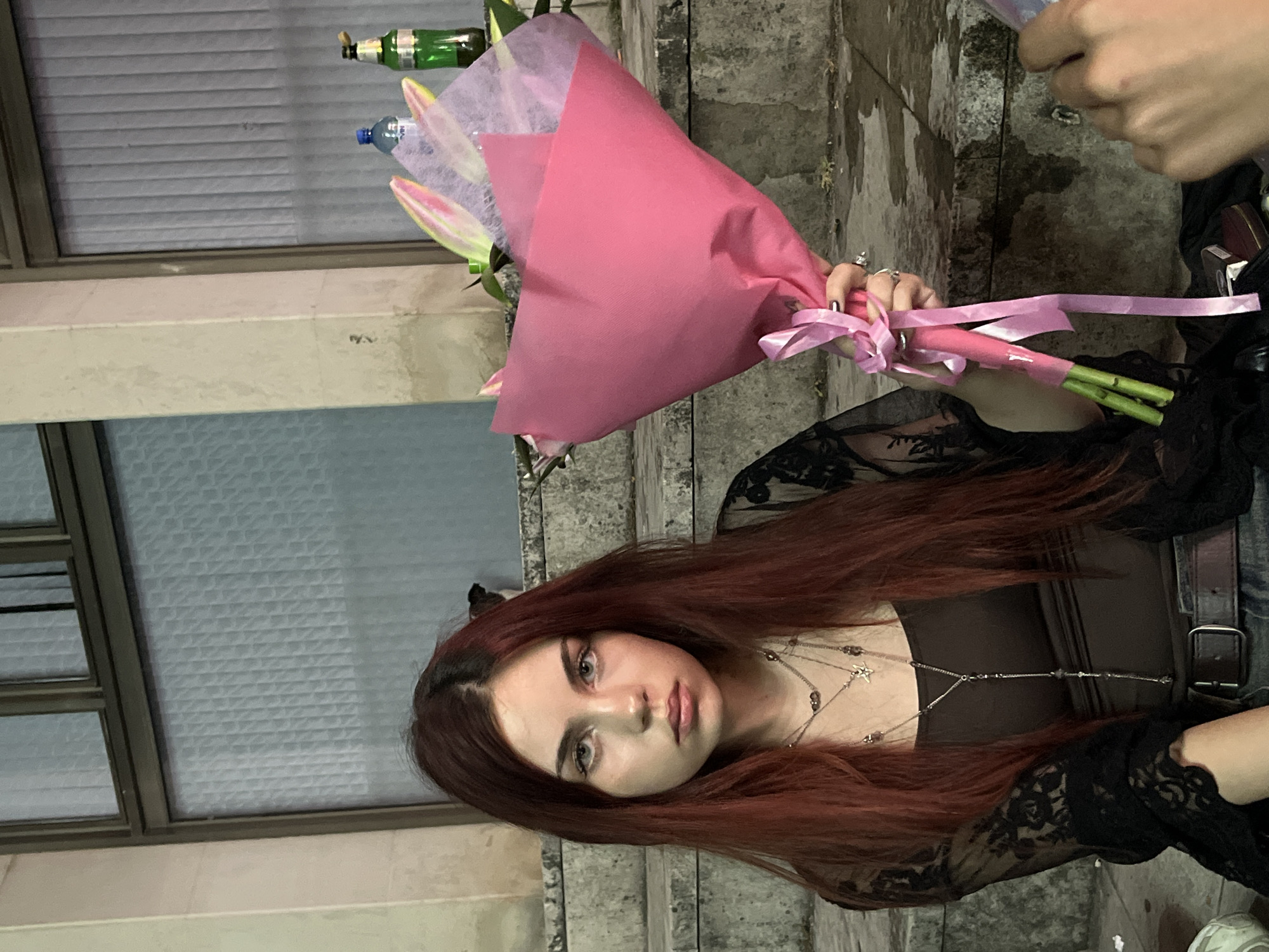 A young woman with long, reddish-brown hair sits on stone steps, holding a bouquet of pink flowers wrapped in bright pink paper and tied with a pink ribbon. She is wearing a dark brown tank top, a black lace overlay top, a belt, and jeans. The woman has a neutral expression, looking directly at the camera. Her neck is adorned with layered silver necklaces, one of which has a star pendant. The background consists of large stone steps and a building with large windows and vertical blinds. To the right of the woman, a bottle of water and a green bottle are visible on a step. Another person's hands are visible in the lower right corner of the image, suggesting they are holding or interacting with the bouquet. The setting appears to be an outdoor public space, possibly near a building in the city of Vratsa, Bulgaria. The stone steps are wet in places, indicating recent rain.