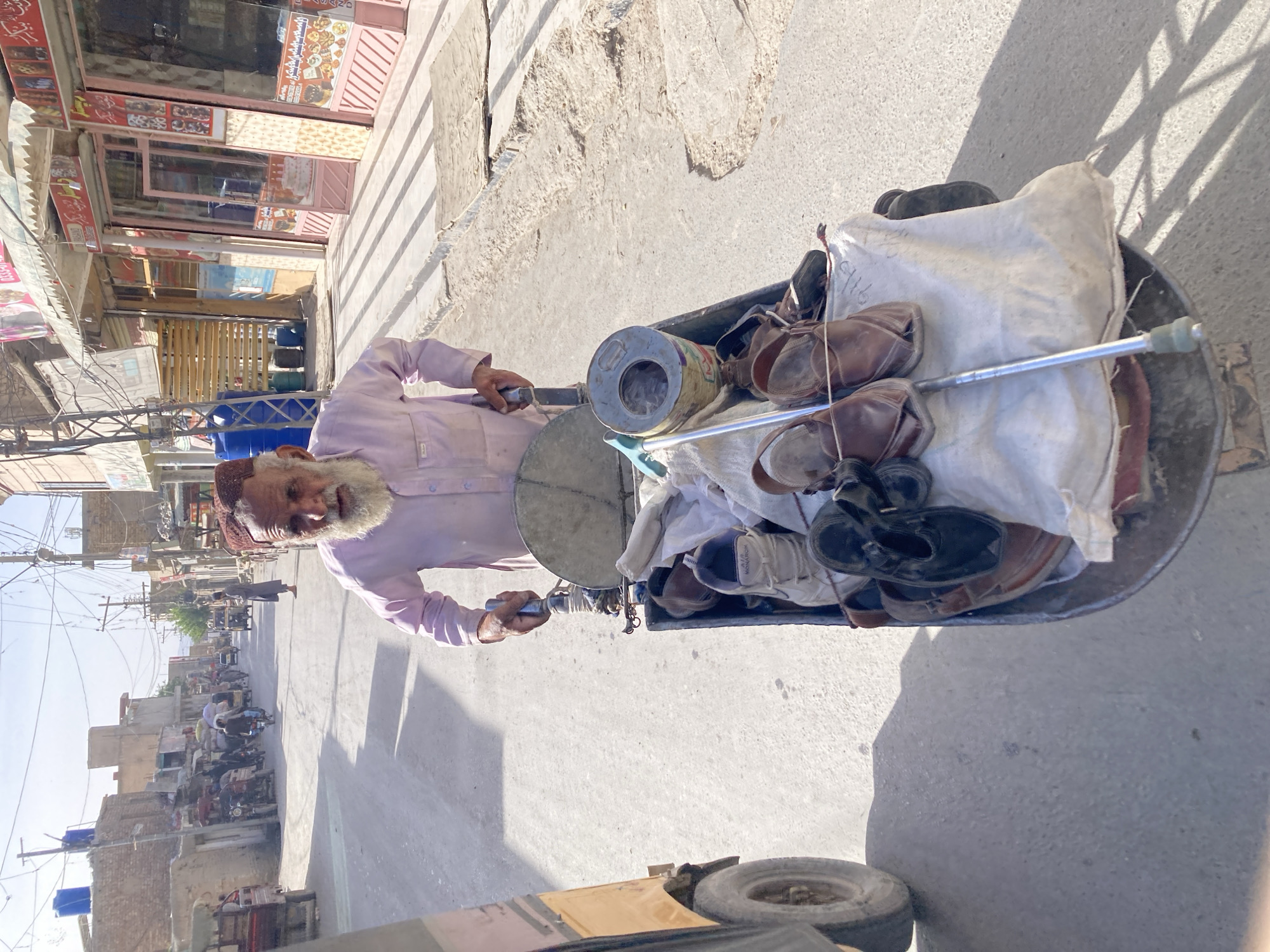 The image shows an elderly man with a white beard and a maroon cap pushing a cart loaded with shoes and other items down a street in Quetta City Tehsil, Pakistan. The man is wearing a light purple shirt. The cart contains an assortment of used shoes, a white bag, a metal container, and a few poles. The street is paved and lined with buildings, some of which appear to be shops. There are motorcycles and people in the background, suggesting a moderately busy street scene. The lighting indicates it is likely daytime.