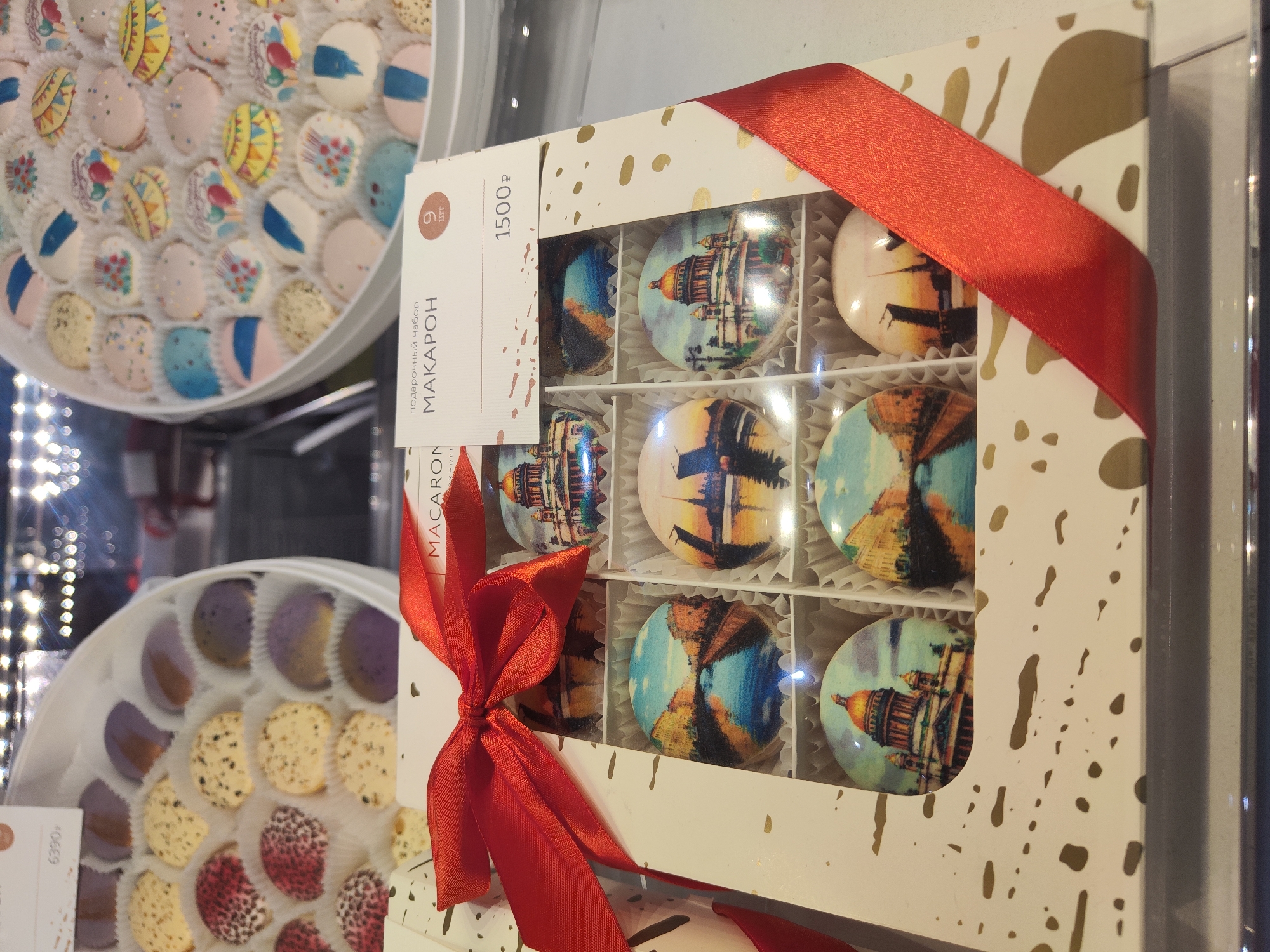 The image captures a retail display featuring several boxes of macarons, likely in a confectionery or gift shop.
The central and most prominent object is a white rectangular gift box tied with a bright red satin ribbon, positioned vertically. This box has a transparent window revealing nine macarons, arranged in a 3x3 grid. Each macaron inside this box is adorned with an edible print depicting a landmark or cityscape. Several macarons clearly display images of Saint Isaac's Cathedral, identifiable by its distinctive golden dome, suggesting the location is Saint Petersburg, Russia. Other macarons feature scenic views, possibly of the city's canals or other architectural elements. The white box itself is decorated with irregular gold splatter patterns.
A white label attached to the central gift box displays Cyrillic text. Visible words include "подарочный набор" (gift set), "МАКАРОН" (macaron), "9 ШТ" (9 pieces), and a price of "1500 ₽" (1500 rubles).
To the upper left, a portion of a large, round white box is visible, filled with numerous macarons in diverse colors and patterns, including blue and white stripes, pastel solids, and various speckled designs. Below this, on the lower left, another large, round white box contains macarons with distinct designs: some feature a purple-to-brown gradient, others are speckled yellow, and a third type is speckled red. A price tag showing "6390 Р" is partially visible near this box.
The scene depicts a vibrant presentation of confectionery items, likely intended for sale as gifts or premium treats. The lighting is bright, creating reflections on the transparent surfaces. No people are visible in the image.