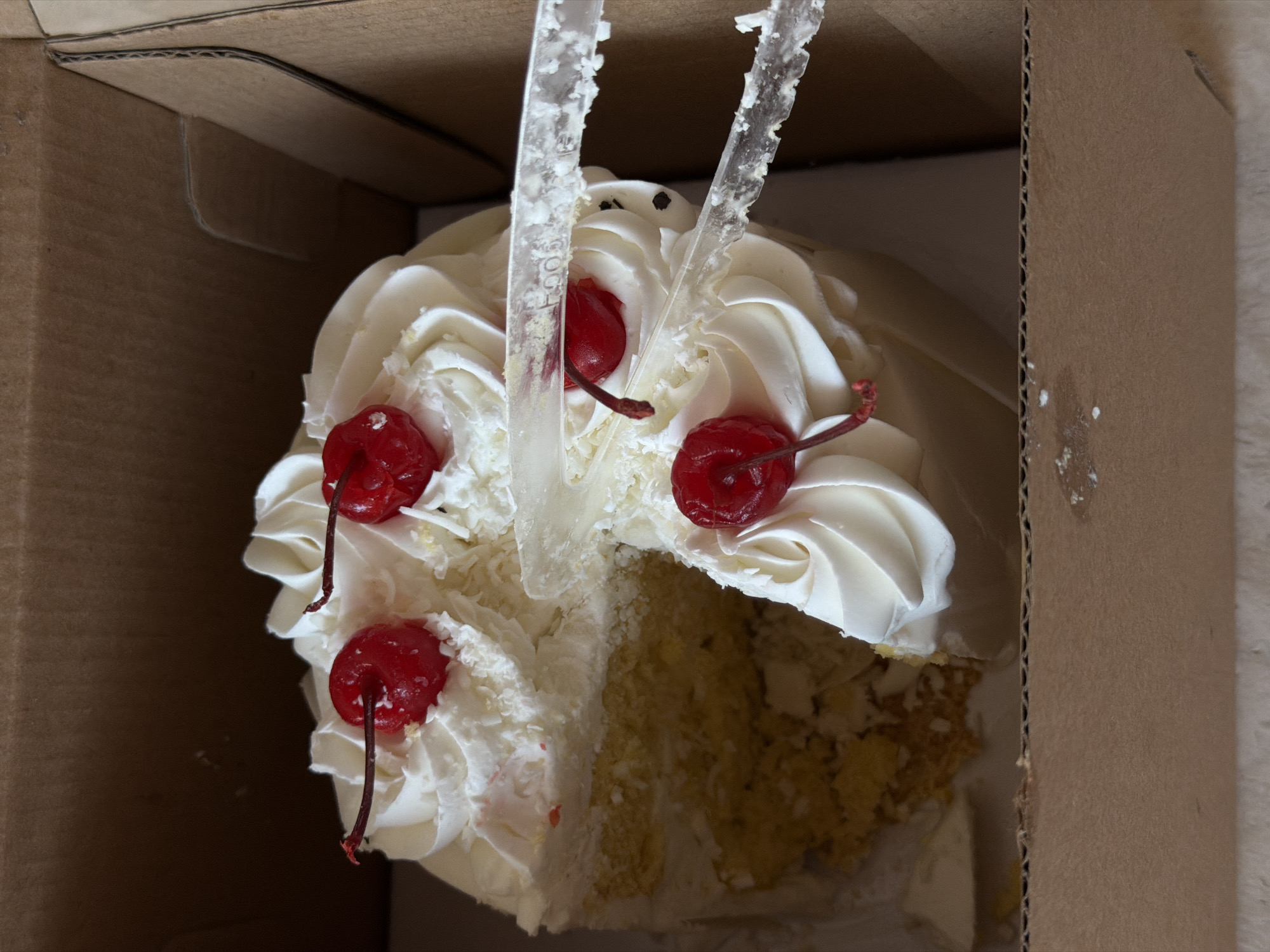 A partially eaten white cake with white frosting and maraschino cherries sits inside a cardboard box. Two clear plastic cake servers with serrated edges are positioned on top of the cake, one on top of the other. The cake is moist and appears to be yellow or vanilla flavored. The box is open and its sides are visible. The image shows the cake from a top-down perspective. There are no people or other living beings visible in the image. The environment appears to be indoors, with a neutral, non-descript background. The location in Mengwi, Indonesia is not discernible from the image.