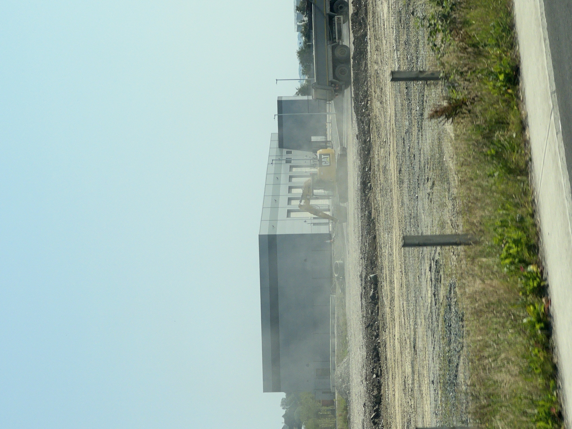 This is a photograph taken on a sunny day in the city of Letterkenny, Ireland. The image captures a construction site with a large, modern, grey building in the background. In the mid-ground, a yellow excavator, identified by "CAT" branding, is actively working, with dust clouds indicating recent activity. Adjacent to the excavator is a large, grey dump truck with multiple wheels, also appearing to be part of the construction operation. The foreground of the image shows a rough, unpaved ground with scattered debris, suggesting ongoing earthmoving and preparation for further construction. A low fence made of vertical posts and horizontal wire mesh runs across the mid-foreground. Beyond the fence, tall green grass and vegetation are visible. The sky is clear and pale blue. The scene depicts a typical industrial or commercial construction project in progress.