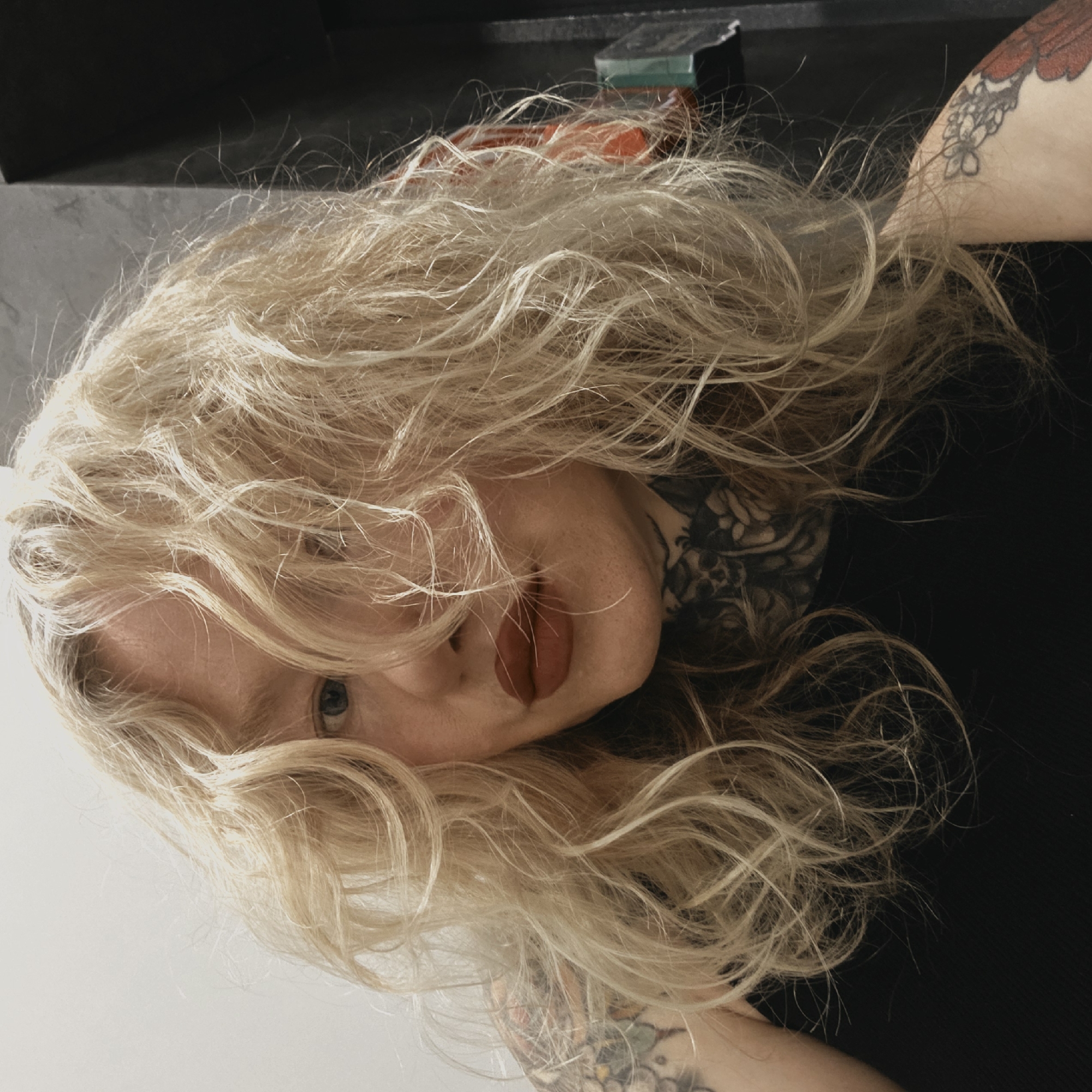 A selfie-style close-up photograph features a person with light blonde, wavy hair partially obscuring their face. The person has fair skin, one visible eye that appears to be blue, and lips painted with a dark red lipstick. The lower portion of their face is visible, including the nose and mouth.
The person has tattoos on their arms and neck. The background is mostly dark, with a few indistinct objects on a surface behind the person. The image appears to be taken indoors with artificial lighting. The photograph is oriented in a tilted perspective, with the person's head appearing at an angle.
The location in Saint Petersburg, Russia, cannot be determined from the visual elements in the image. There is no way to confirm the location as Saint Petersburg.
