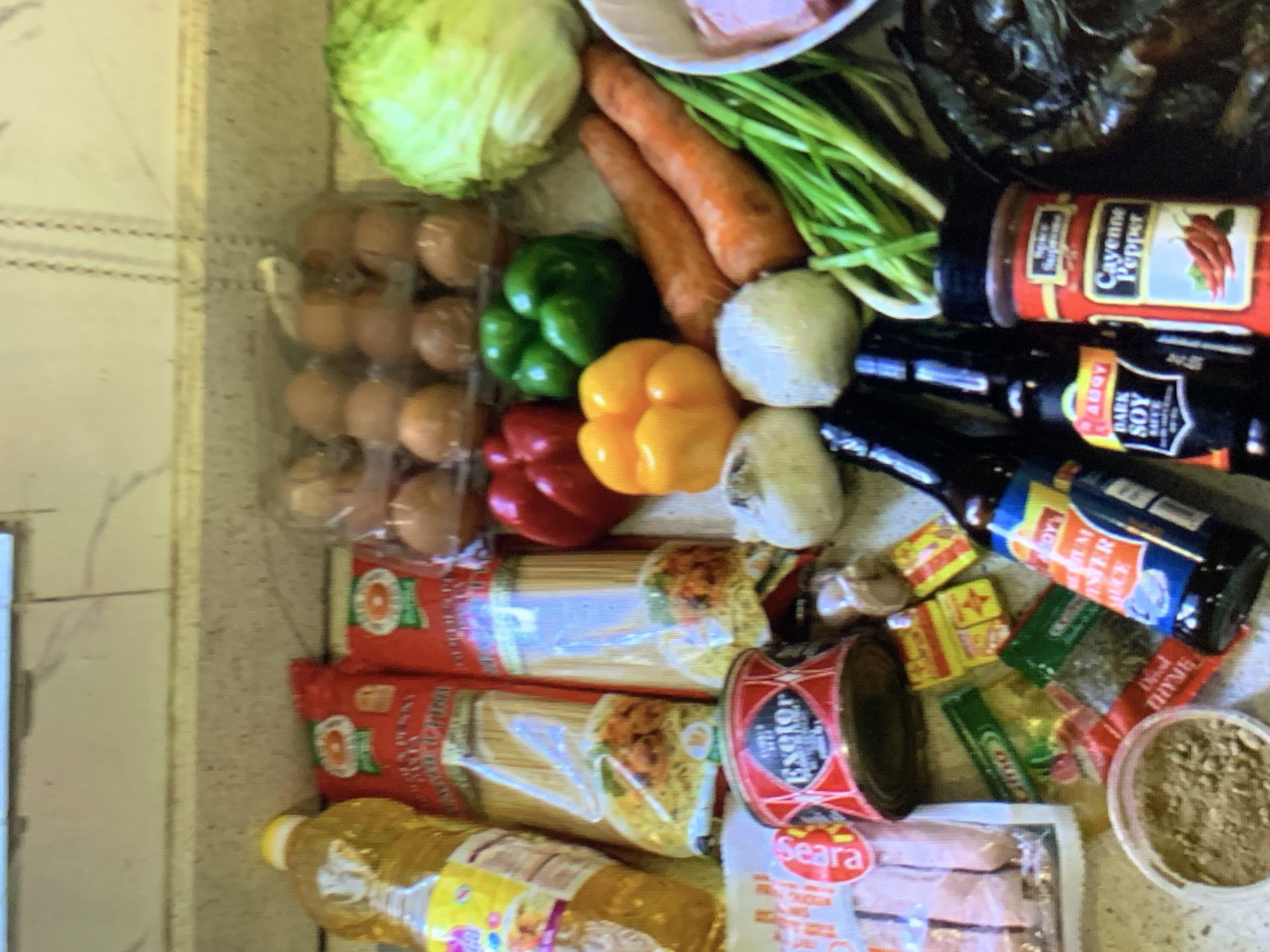 The image shows a countertop with various ingredients. A head of lettuce, a container of eggs, carrots, green onions, and green, yellow, and red bell peppers are present. There is a can of sardines and packages of spaghetti. Spices include cayenne pepper and dark soy sauce. A bottle of cooking oil and other smaller packets of spices and seasonings are also visible.
The location in Rafin Par, Nigeria, is assumed from the query and cannot be verified from the image itself. There are no people visible in the image.