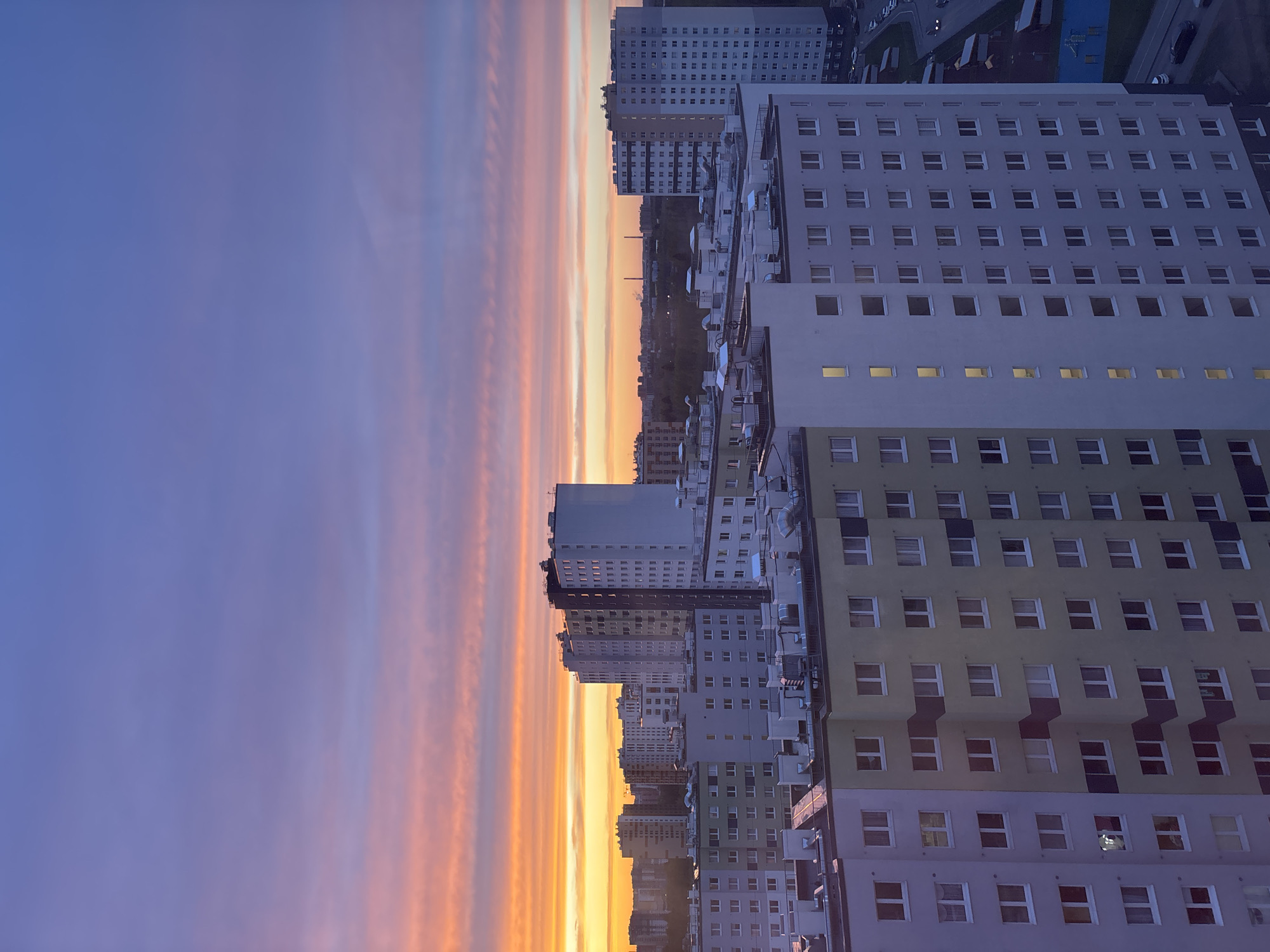 The image shows a cityscape at dusk or dawn. The sky has a gradient of colors, transitioning from a deep blue to orange and yellow near the horizon. Several tall apartment buildings are visible, with multiple rows of windows. The buildings range in color from white and gray to a light green. Some of the windows are lit, suggesting activity inside. The scene appears to be in a densely populated urban area.