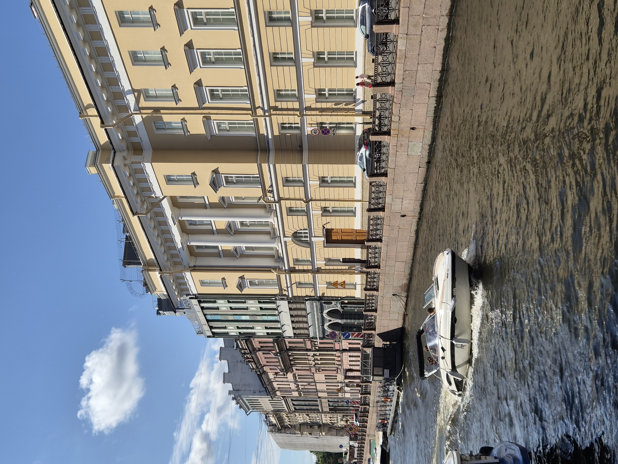 In a European city, a white speed boat with three occupants navigates a wide canal lined by buildings. The boat, with a black canopy and the registration "GER 4649," is leaving a wake. On the right bank, a neoclassical yellow building with white columns and numerous windows stands prominently, featuring a decorative cornice and ornate railings along the canal. Adjacent to it are a series of multi-story buildings with diverse architectural styles and colors, including pink, gray, and green. The canal's edge is fortified with a stone embankment. On the sidewalk behind the railings, two individuals are walking, and two cars are parked. The sky is blue with scattered white clouds.