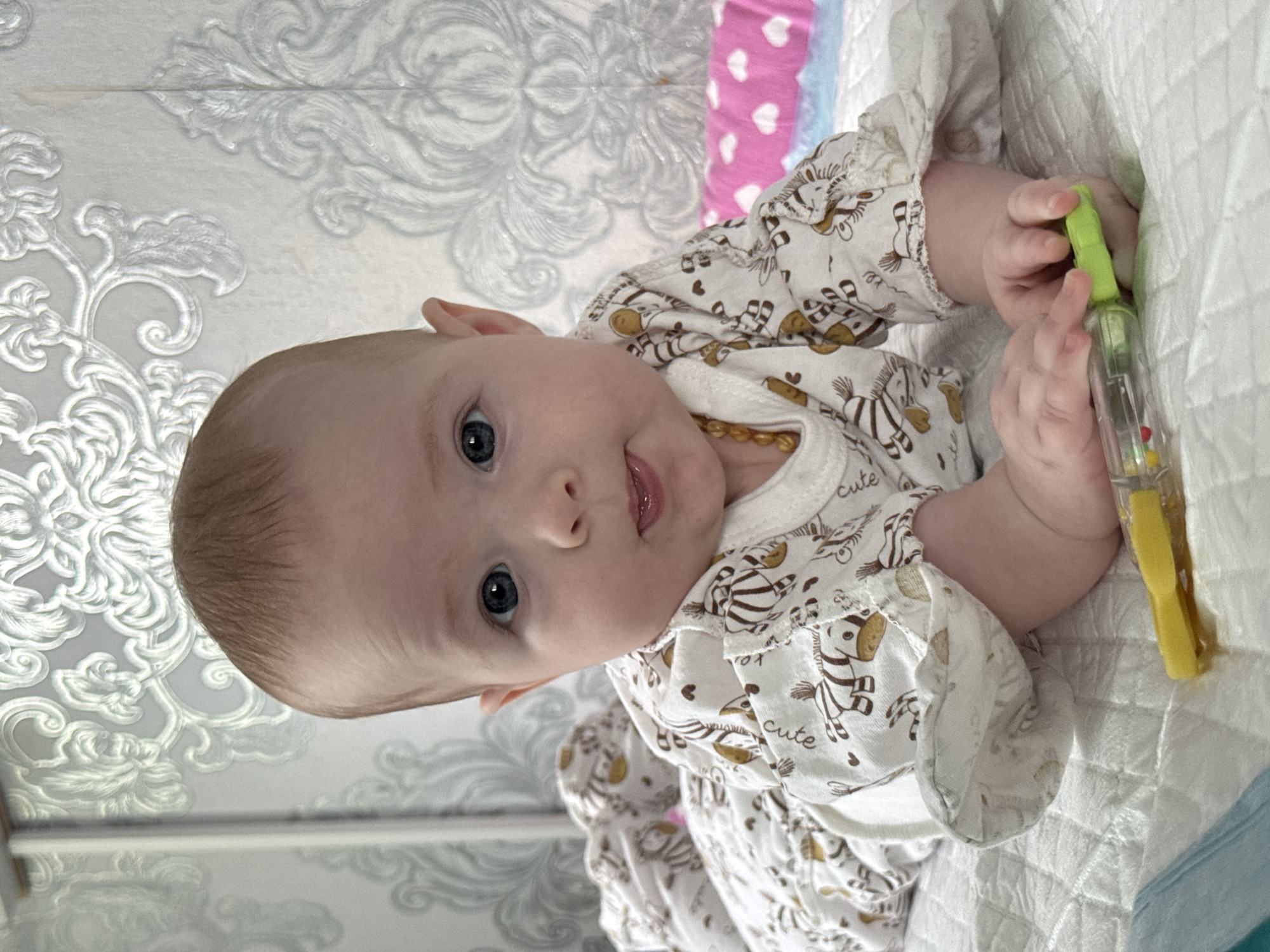 A baby with blue eyes, wearing a white outfit with zebra print and the word "cute" repeated, is lying on a white surface with a pink and white patterned fabric in the background, possibly in the city of Кемуль, Russia. The baby is holding a green and yellow toy in its hands. The background includes a wall with a patterned design. The scene appears to be indoors.