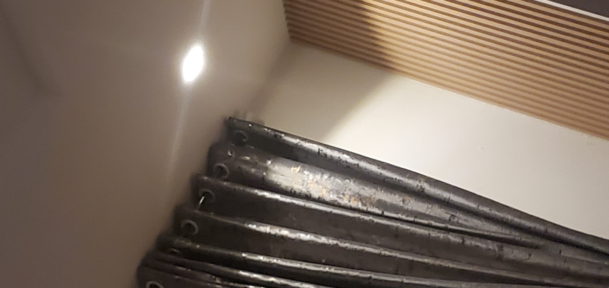 This is a close-up, low-angle shot of a stack of dark, cylindrical objects, possibly tent poles or structural supports. They are piled on top of each other, with the uppermost poles appearing worn and scratched, showing hints of lighter material underneath. The poles are arranged in a way that suggests they are stored in a corner, with a plain, light-colored wall curving away to the left. A bright, circular light source is reflecting off this wall, creating a soft glow. In the upper right quadrant of the image, a section of a wall with horizontal wooden slats is visible, suggesting an interior setting. The overall lighting is dim, with the primary light coming from the reflection on the wall. There is no discernible activity taking place, and no people or text are visible. The context provided places this scene in Gura Top, Nigeria, which does not offer specific visual cues within the image itself, but implies a potential outdoor or storage context for the poles.