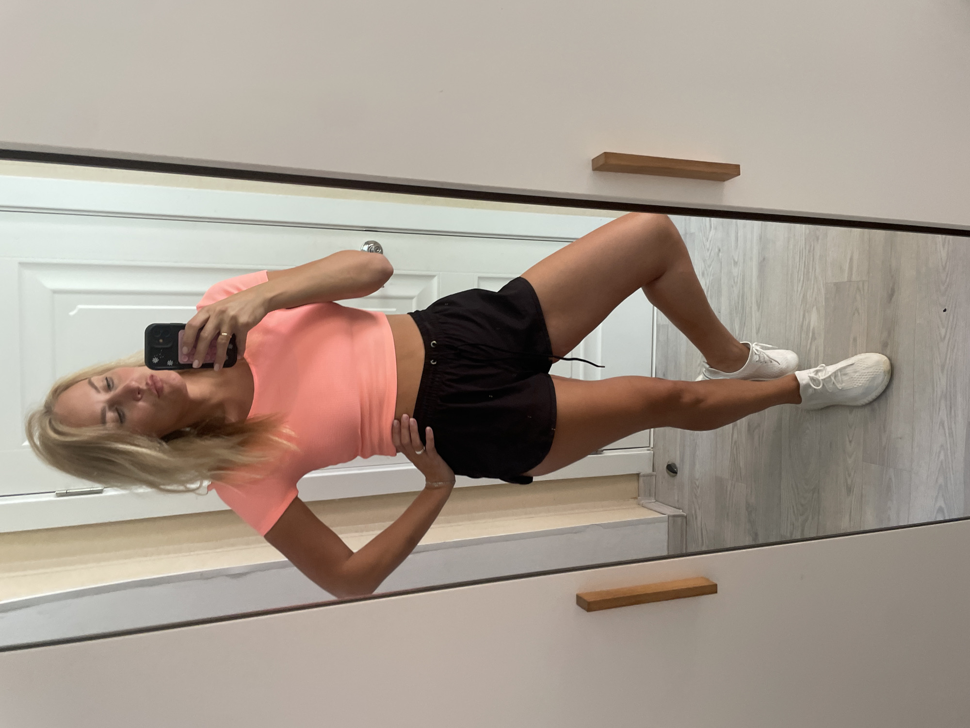 A woman with blonde hair is taking a selfie in a mirror. She is wearing a coral crop top, black shorts, and white sneakers. She is standing in what appears to be a bedroom or dressing area. The room has light wood flooring and a white door in the background. There are two closet doors visible, one with a wooden handle. The woman is posing with one leg bent at the knee. The image is taken in Verkhniy Yurt, Russia.
