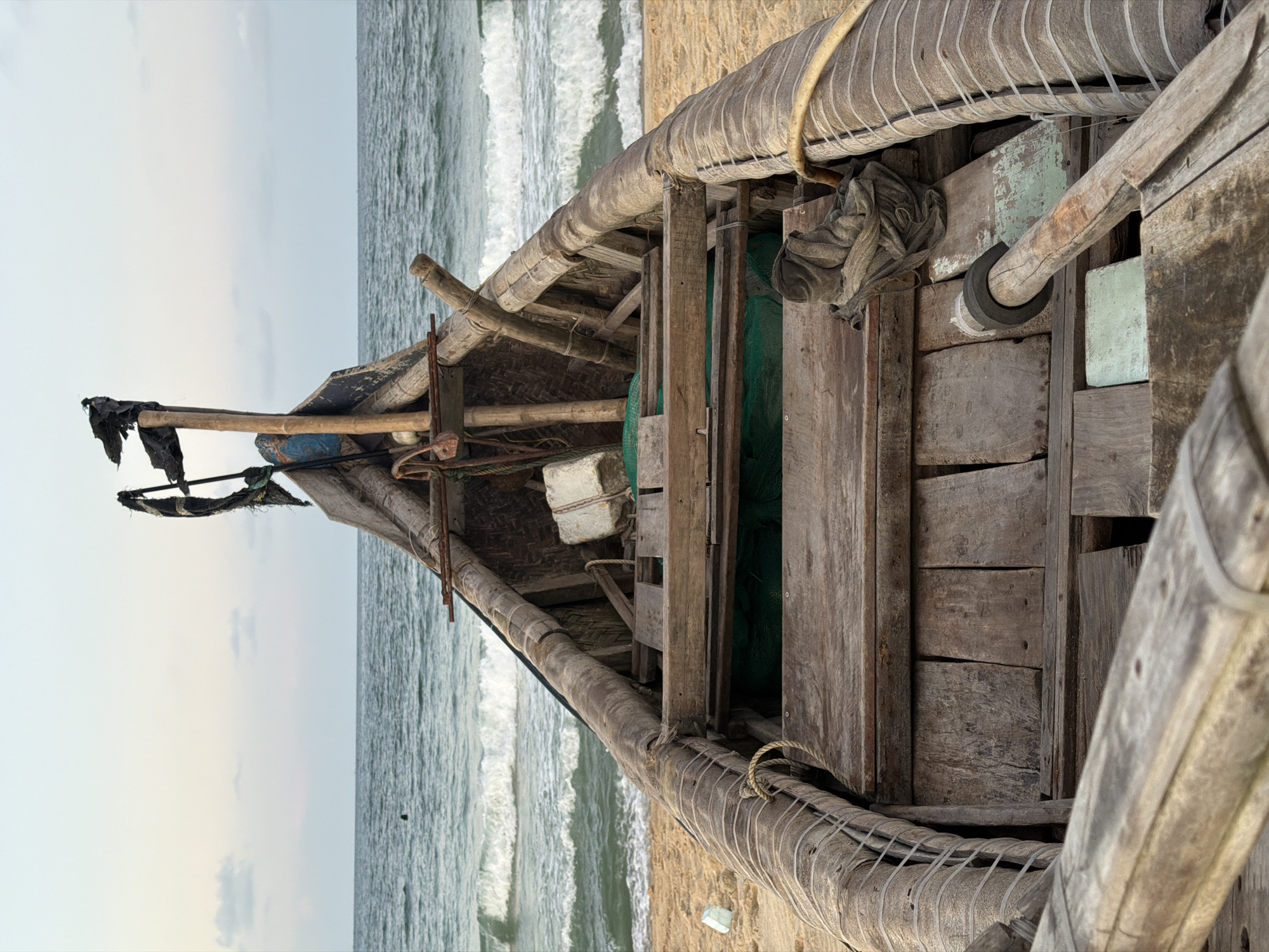 This is an outdoor shot of a rustic, wooden boat on the sandy shore of Thuận An Ward, Vietnam, facing the ocean. The frame is oriented vertically, showing the bow of the boat pointed towards the horizon. The boat is weathered, constructed from roughly hewn wooden planks and beams. It is fitted with various ropes, netting, and other implements, indicating it is a fishing vessel. The ocean's water is churning with small waves. The sky above the horizon is a pale, overcast gray.