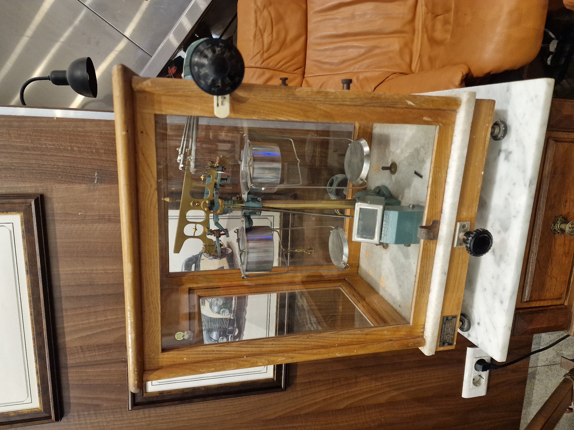 The image shows an antique balance scale housed in a wooden and glass display case. The scale itself is intricate, with polished metal components and two circular pans. It is positioned on a marble surface, which sits atop a wooden cabinet. To the left of the display case, a framed picture with a light-colored mat and dark border is visible. The background includes a portion of a brown leather sofa and a black adjustable lamp. An electrical outlet with a black plug is also visible at the bottom right. The scene appears to be indoors, likely in a room with wooden furniture and a marble accent. There is no activity or event depicted; the image is a still life of the object. Some text is visible on a small metal plate attached to the side of the display case, but it is too blurry to read.