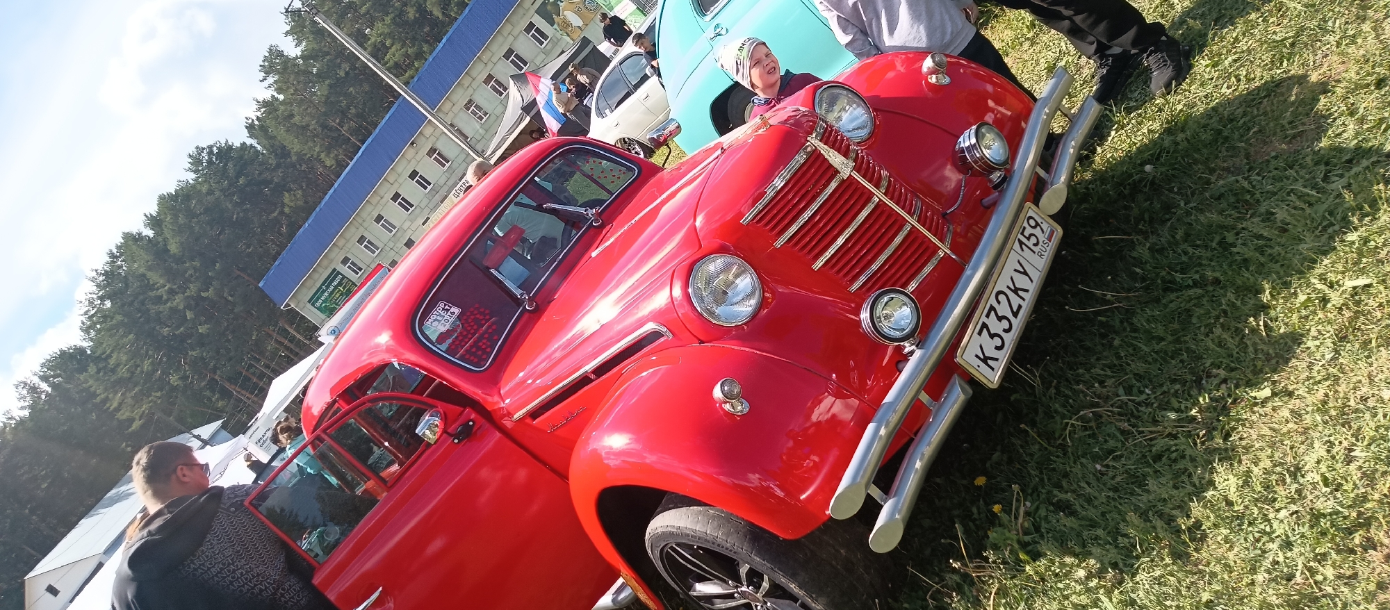 The scene shows a red vintage car, possibly a modified Moskvich 400 or 401, parked on a grassy surface. A person wearing a hat and a dark-colored top is standing near the front of the car. A number plate is visible, reading "K332KY 159 RUS". Behind the car, there are other vehicles and people in the background, suggesting an outdoor event or gathering. A building with a blue roof and white windows is visible in the background. The environment indicates a sunny day. The location appears to be a public space.