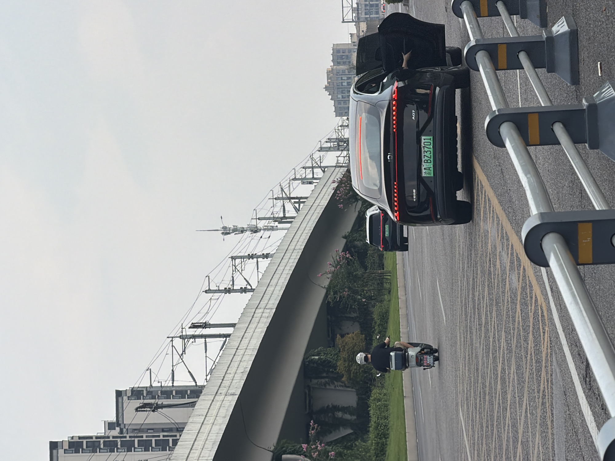 The image depicts a street scene in Jiangjin, China, with a focus on traffic and urban infrastructure. A black electric vehicle, identified by its license plate "A BZ3701", is prominent in the foreground, with its driver's side door open and a person partially visible inside. To the left, a person on a white electric scooter rides on the road, wearing a black shirt and shorts, and a white helmet. In the background, a multi-level overpass, likely for a railway or highway, dominates the upper portion of the frame, supported by concrete pillars. Overhead power lines for an electrified railway are clearly visible, stretching across the scene. The environment is characterized by a gray, overcast sky and urban elements including residential buildings with multiple windows and telecommunication towers. The road surface is asphalt, marked with yellow and white lines. Safety barriers with yellow accents are positioned in the foreground, separating lanes of traffic. Greenery, including bushes and vines, grows along the roadside and on the supports of the overpass, suggesting a blend of urban development and landscaping. Another car with license plate "A 091537" is visible further down the road, also traveling in the same direction. The overall impression is one of a typical urban environment in motion.