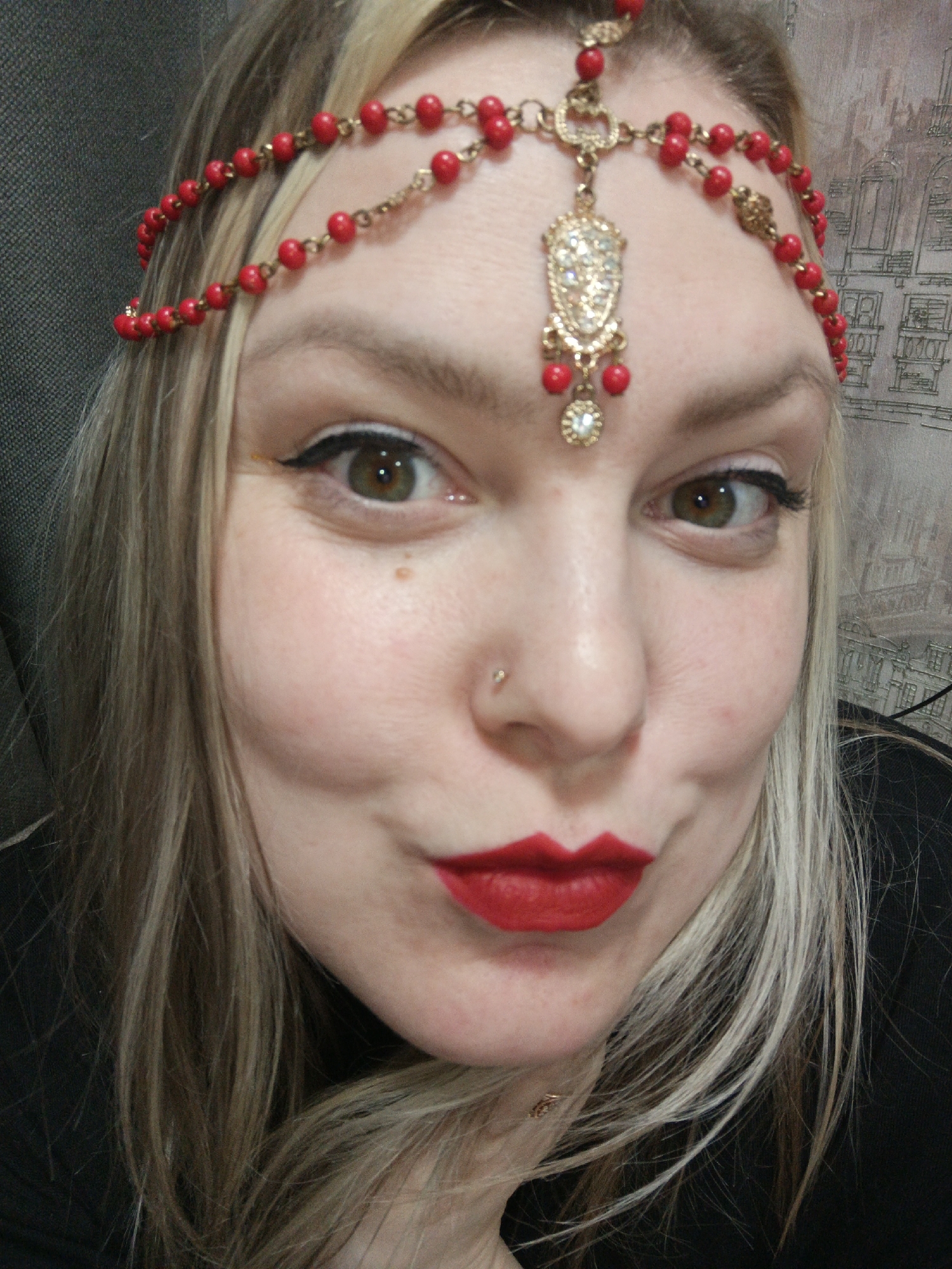 A close-up shot of a woman's face with fair skin, light brown hair with blonde highlights, and green eyes with black eyeliner. She has red lipstick on and is wearing a red and gold beaded headpiece with a decorative pendant in the center of her forehead. She has a small stud piercing in her left nostril. The background appears to be a gray textured wall and a patterned wallpaper. The city of Марковский, Russia cannot be determined from this image.