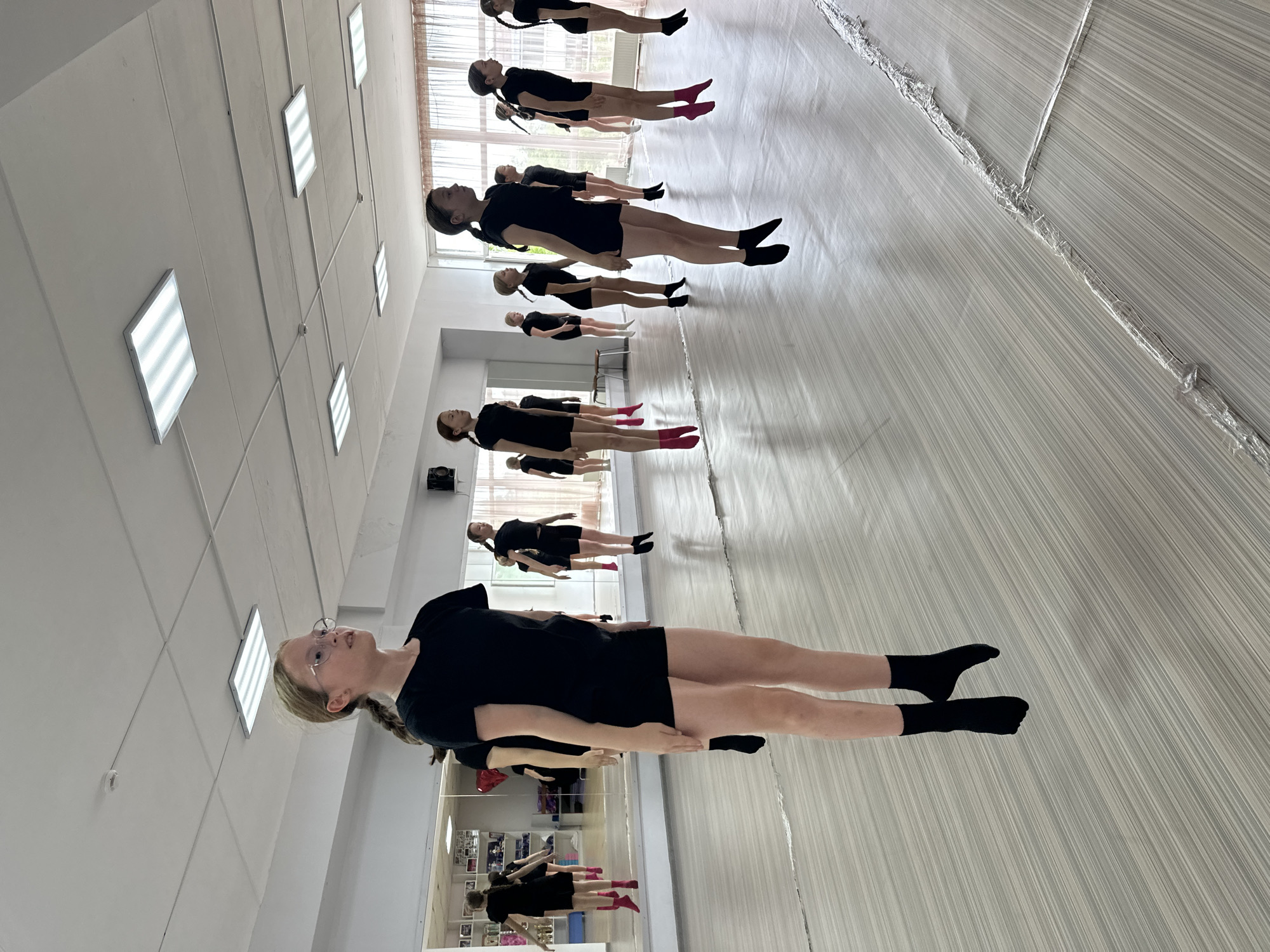 In Votkinsk, Russia, a group of young girls, all wearing black leotards and shorts, are practicing a dance routine in a studio. They are in mid-air, appearing to be jumping or levitating. The studio has a mirrored wall on the left side and a large window with curtains on the right. The floor is a light-colored wood or laminate with a striped pattern. The lighting fixtures on the ceiling are rectangular and fluorescent. The girls are positioned at various depths in the frame, with some closer to the camera and others further away, creating a sense of depth and movement. The girl in the foreground, on the left, is wearing glasses and has her hair in a braid. Some of the girls are wearing colored socks, either pink or black. The overall atmosphere suggests a dance class or rehearsal.