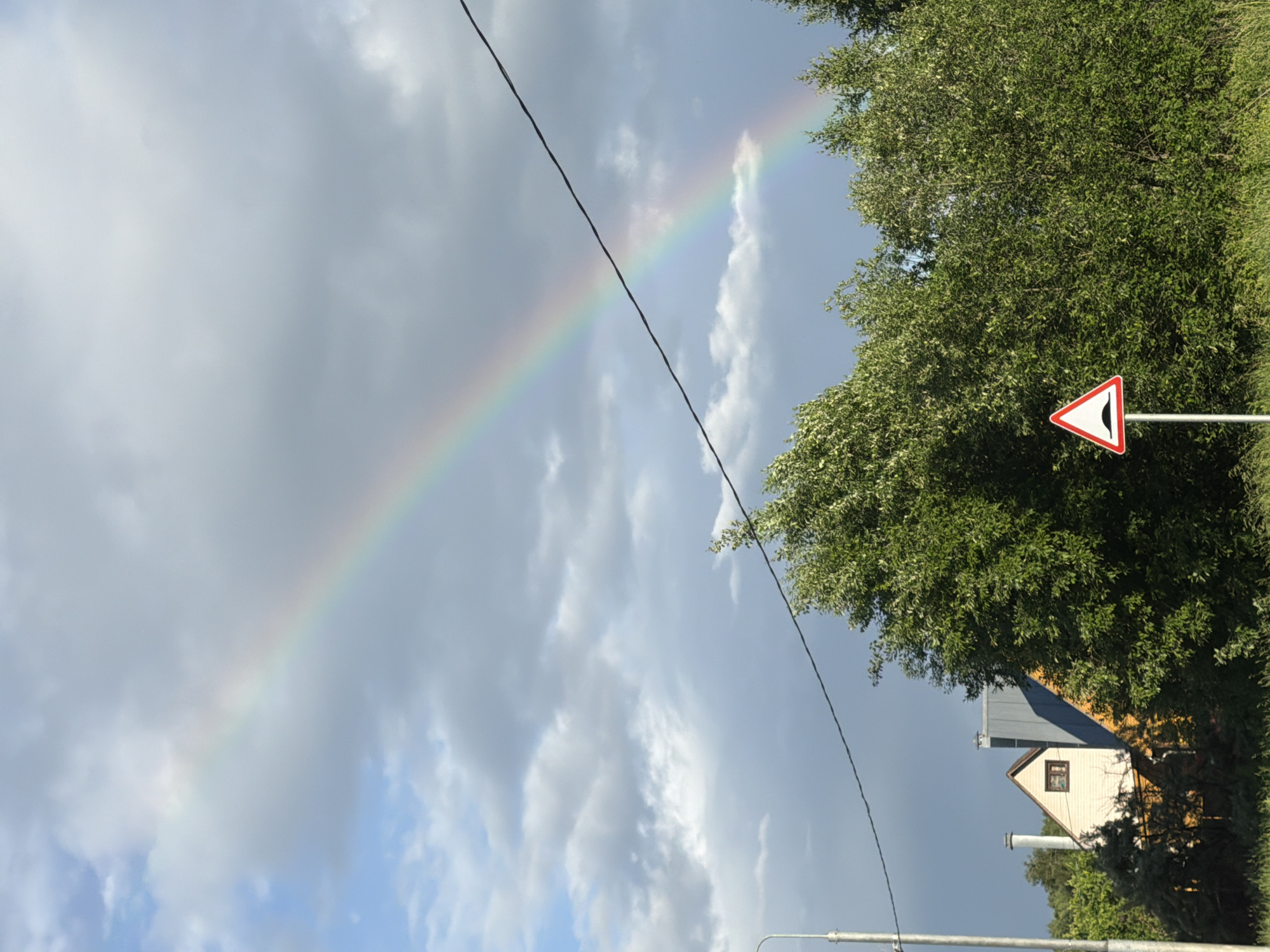The image captures a view of a suburban landscape with a rainbow arcing across a cloudy sky. In the foreground, a cluster of lush green trees dominates the scene. To the left of the trees, a white triangular road sign with a red border indicates a speed bump ahead. Behind the trees, a portion of a residential building with a yellow facade and a grey roof is visible. The building appears to be a house, with a small brown-framed window and two chimneys. Further to the left, a light pole stands, and a wire stretches across the upper portion of the frame. The sky is a mix of blue and grey clouds, with the rainbow prominently displayed. The scene suggests a location in Satino-Russkoe, Russia, likely during or shortly after a rainfall. No people are present in the image.