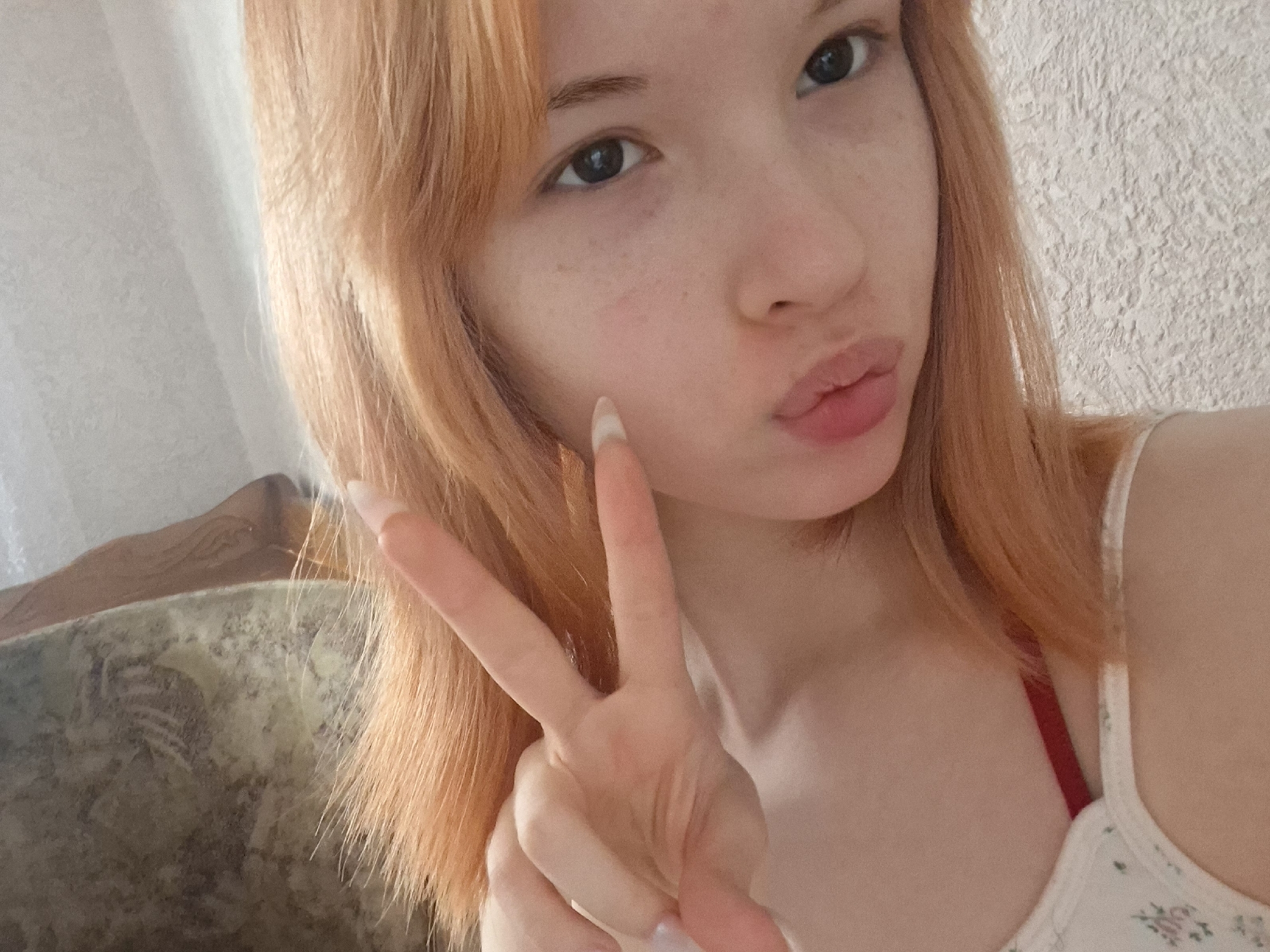 A young woman is taking a selfie, holding up her left hand in a peace sign. She has light orange hair and light skin with freckles. Her lips are pursed. She is wearing a white top with a floral pattern and a red strap. The background includes a patterned piece of furniture. The location is Chaikovsky, Russia.