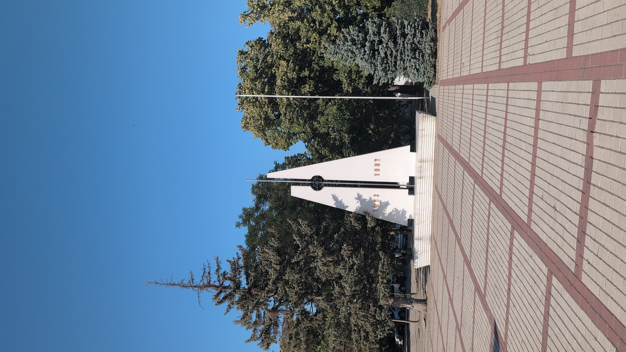 The image shows a monument in the city of Izmail, Ukraine. The monument is a white, triangular structure with a vertical metal rod in the center. Two red numbers, "1923" and "1923", are visible on the sides of the monument. The monument is located in a public square with a paved ground made of rectangular tiles. Several trees, including a tall pine tree on the left and a conical evergreen on the right, surround the monument. The sky is clear and blue, indicating a sunny day. Cars are parked in the background behind the trees on the left. A flag pole stands to the right of the monument. The scene is static, with no people or animals visible.