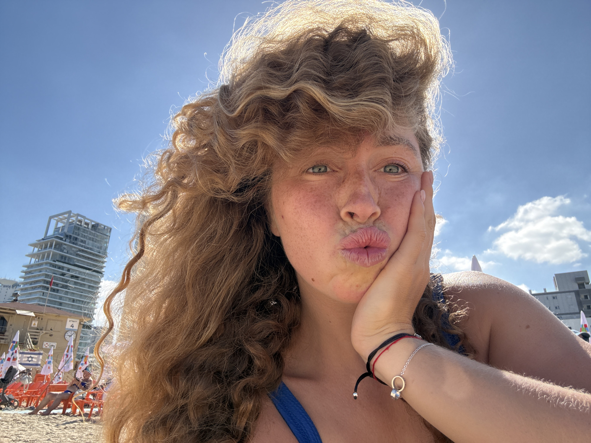 A woman with curly blonde hair and freckles is taking a selfie on a sunny day at a beach in Tel Aviv, Israel. She is wearing a blue swimsuit and has her hand on her cheek, puckering her lips. In the background, there are beach chairs, flags with Israeli symbols, and a modern skyscraper. The sky is clear blue with some clouds.