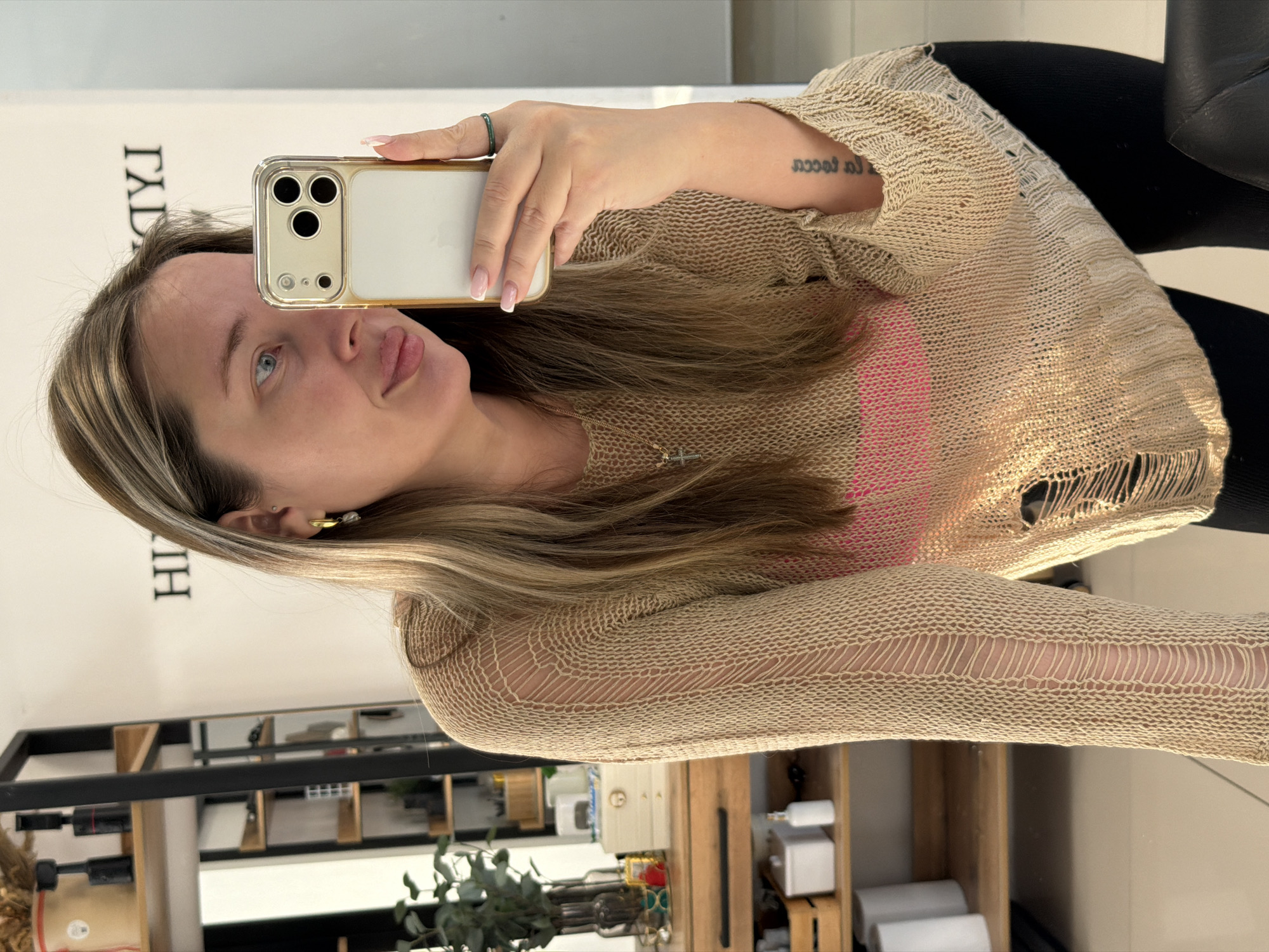 A woman is taking a selfie with an iPhone. She has blonde hair with lighter highlights, blue eyes, and fair skin. She is wearing a loose-knit, beige sweater with distressed details over a pink top. She is wearing a green ring on her left ring finger and has a tattoo on her inner left forearm. She is holding the phone in her right hand with a French manicure. The background includes a partially visible sign with lettering, a black metal shelving unit, and various items on a wooden shelf, including plants and containers. The setting appears to be indoors, possibly a hair salon. There is no explicit indication of the location being Chaikovsky, Russia.