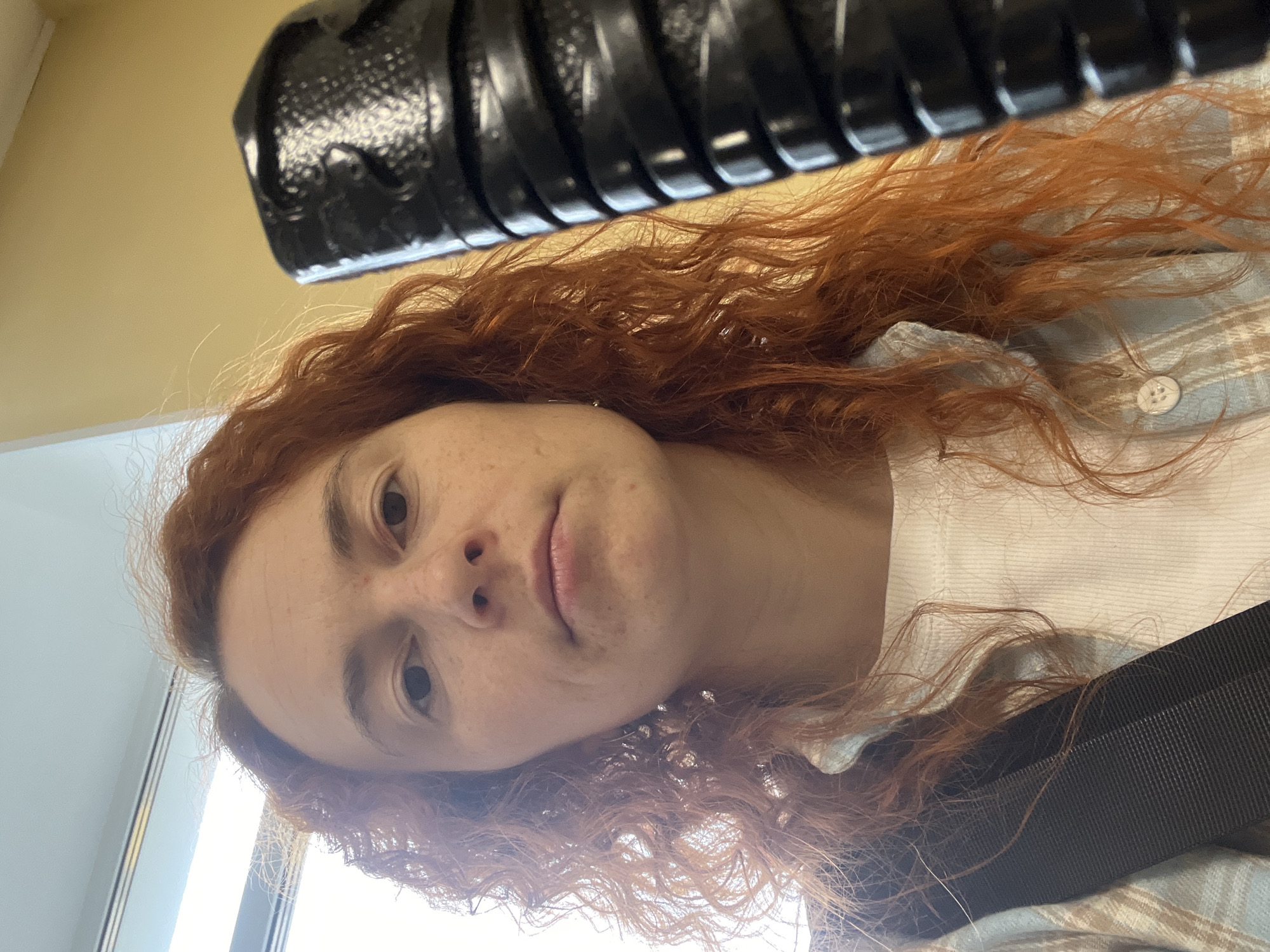 A close-up, rotated image shows a woman with red curly hair looking at the camera. She has fair skin with freckles. She is wearing a white shirt and a light blue plaid button-down shirt. A black corrugated object is above her head. The background appears to be a beige wall and a window. The location is not discernible from the image.