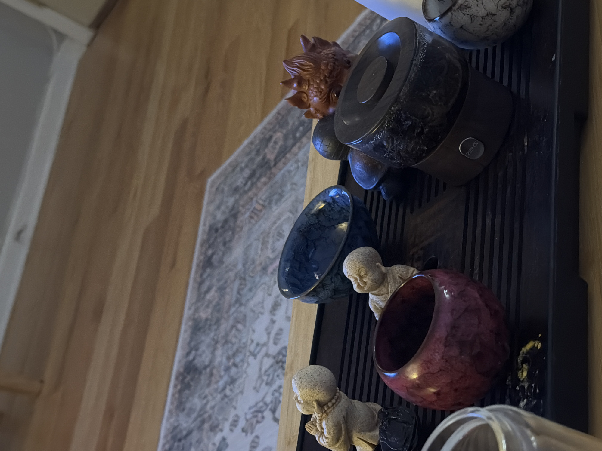 The image shows a collection of Asian-themed objects arranged on a dark, possibly black, slatted surface. The objects include a carved wooden figurine of a lion or mythical creature, a Zensorium Aroma Oil Diffuser with a stone finish, a blue and black bowl, a maroon circular vessel, and two small, pale-colored statues of Buddha-like figures. In the background, a wooden floor is visible on the left, while a patterned area rug is present behind the objects. The scene seems to be indoors. The location of Morton Grove, United States of America, is not discernible from the image.
