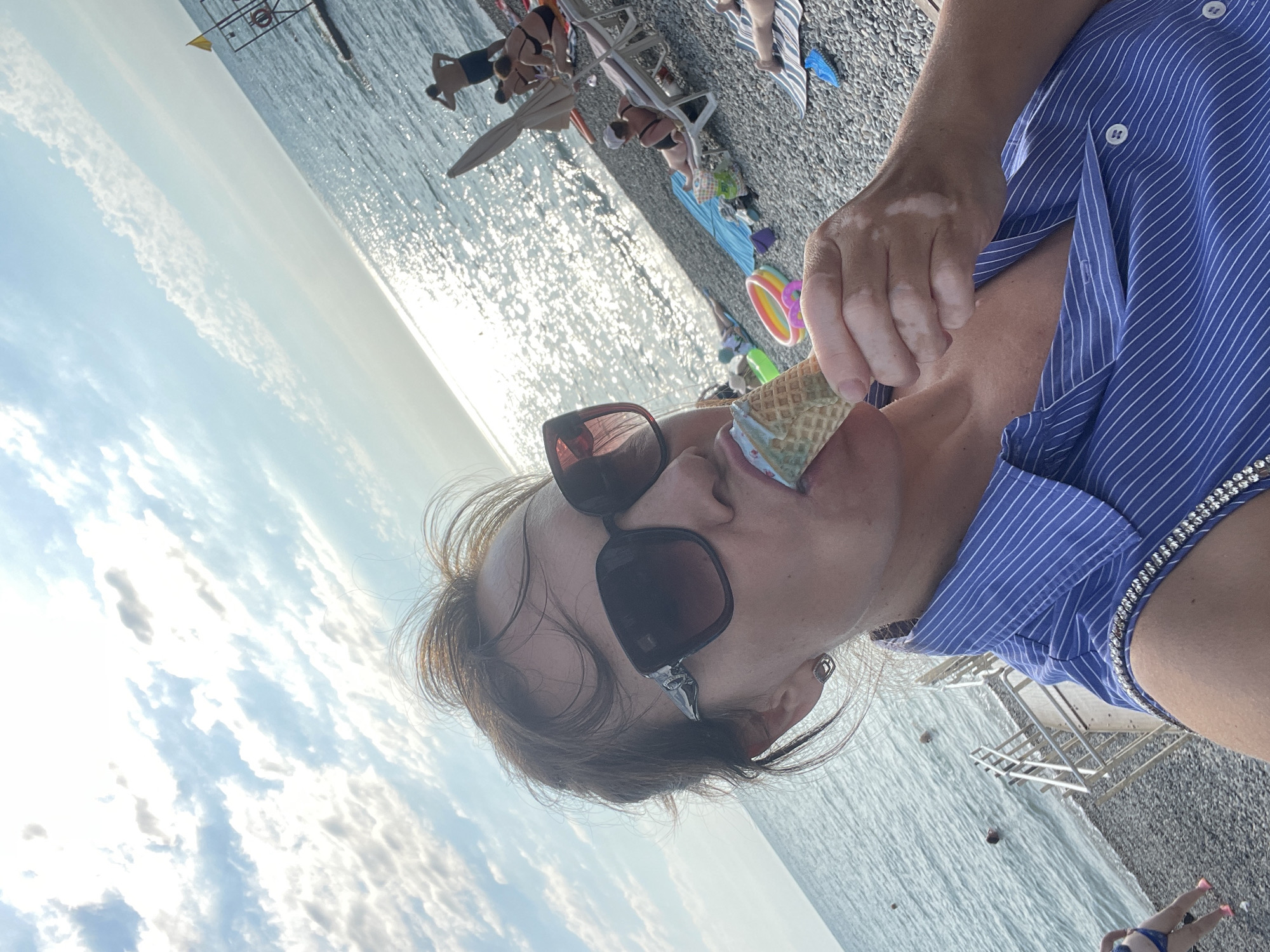 A woman is taking a selfie on a pebble beach in Sochi, Russia, while eating an ice cream cone. She is wearing sunglasses and a blue and white striped shirt. The sun is shining brightly, and the sea is visible in the background, with several people sunbathing and swimming. The sky is partly cloudy.